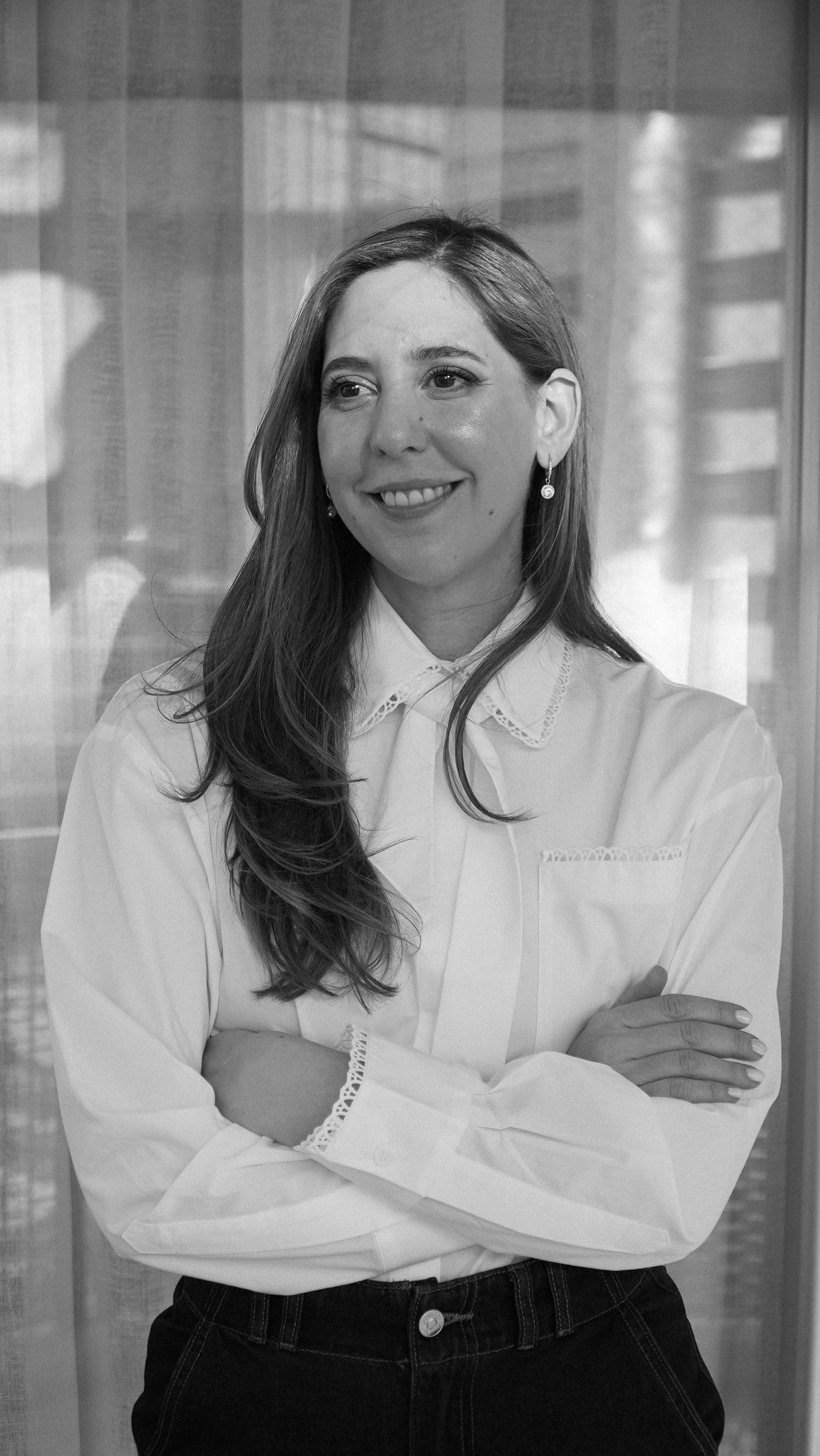 Retrato en blanco y negro de una mujer con cabello largo, sonriendo, con los brazos cruzados, usando una blusa blanca con detalles en el cuello y prendas oscuras, en un ambiente interior.
