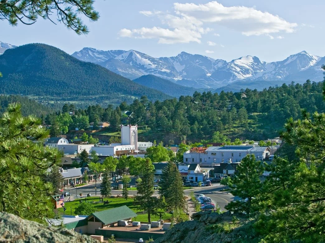 View of a small town surrounded by green trees, with mountains in the background, some with snow on their peaks.