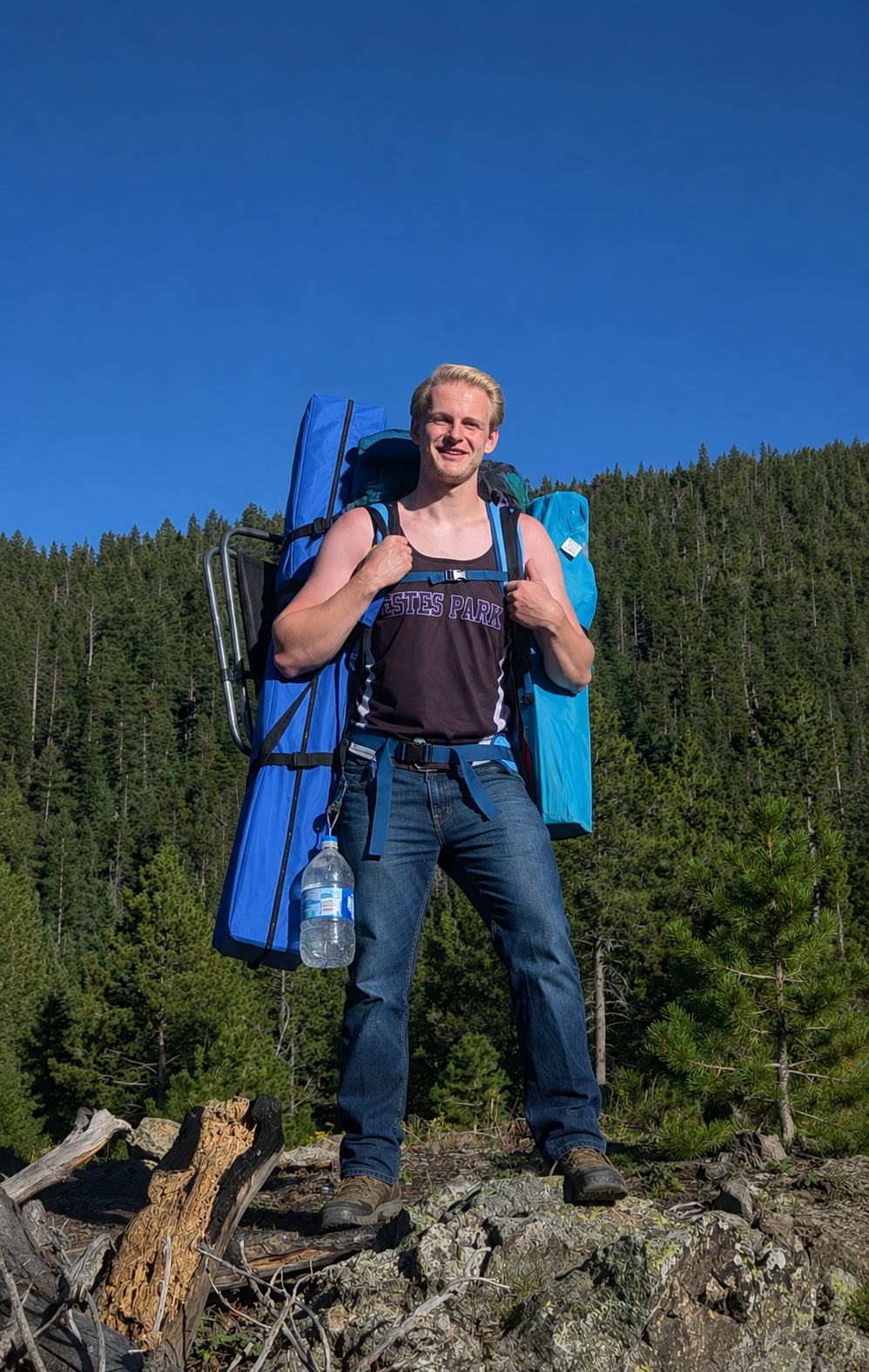 A man standing outdoors on rocky terrain, carrying a large blue backpack with camping gear, in front of a forest with evergreen trees under a clear blue sky.