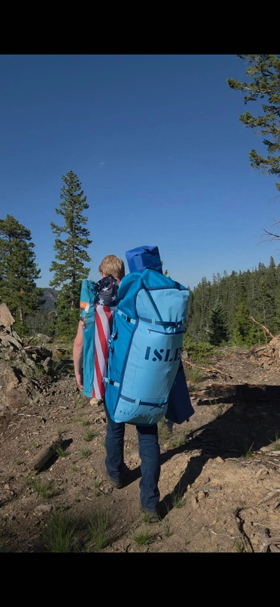 A person hiking in a forest carrying a large blue backpack and wearing a towel with the American flag pattern.