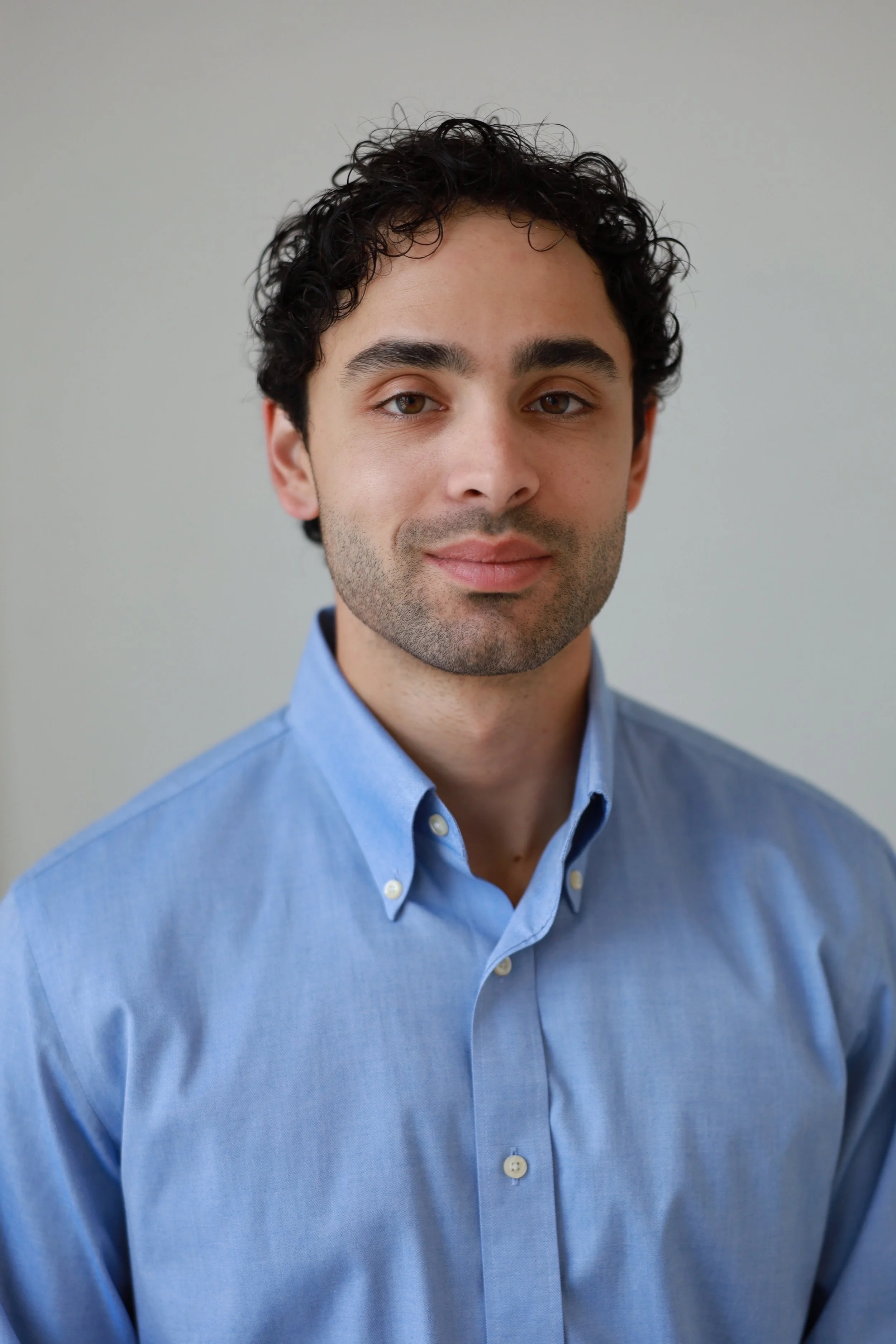 Portrait of a young man with curly dark hair, wearing a light blue button-up shirt, against a plain gray background.
