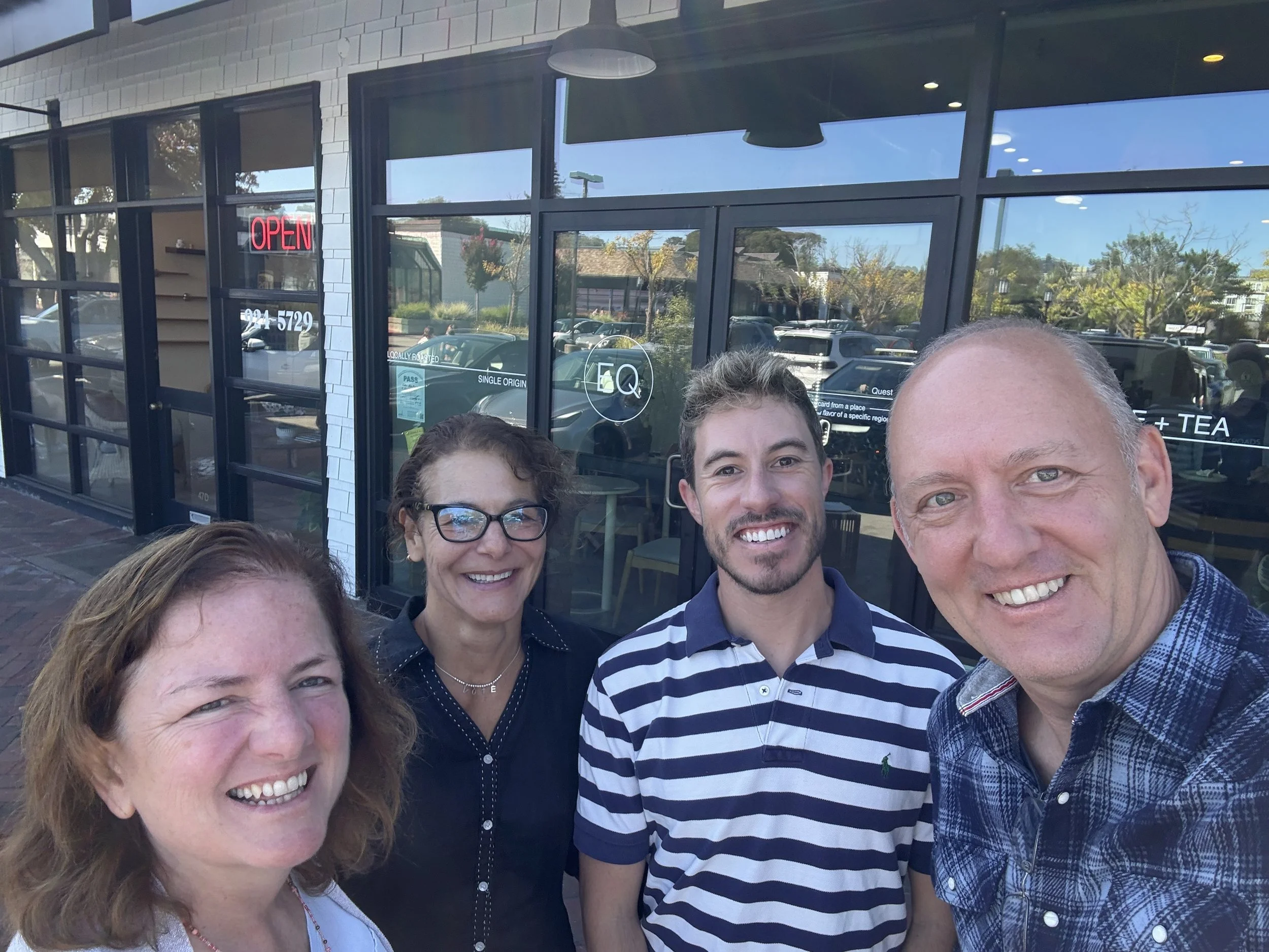 Group of four smiling people taking a selfie outside a glass storefront on a sunny day.