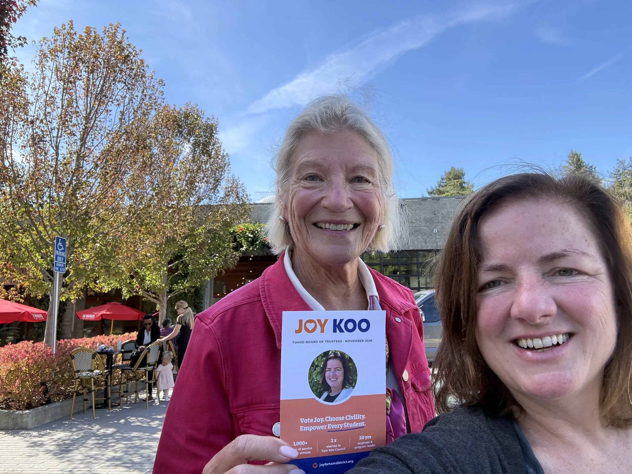 Smiling woman with gray hair holding a political campaign flyer at an outdoor event, with young woman smiling partially visible in the foreground, trees with autumn leaves and blue sky in the background.