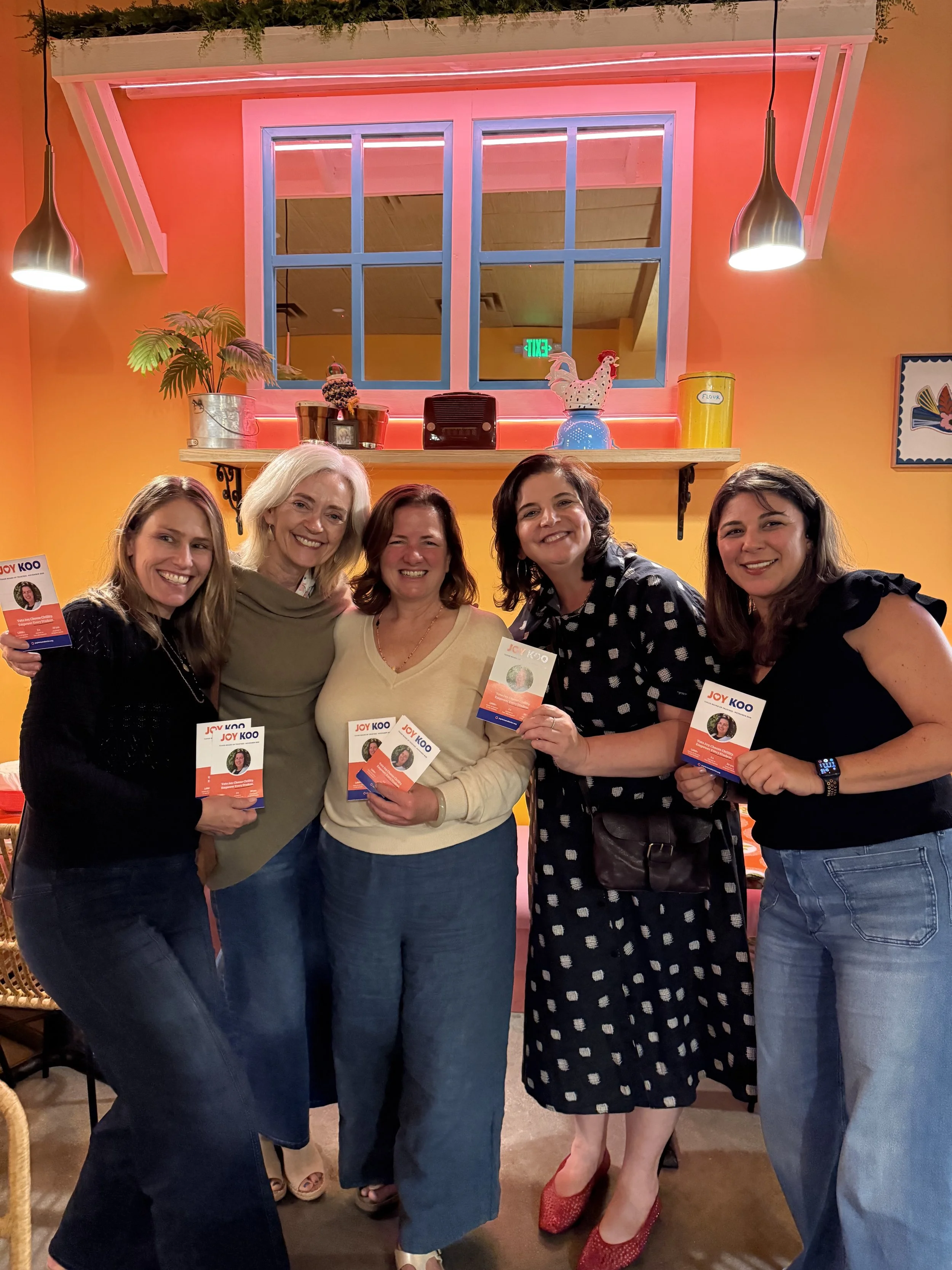 Five women posing indoors, holding flyers with the name 'Joy Koo', standing in front of a vibrant orange wall with decorations including a rooster figurine and a plant, under black pendant lights.