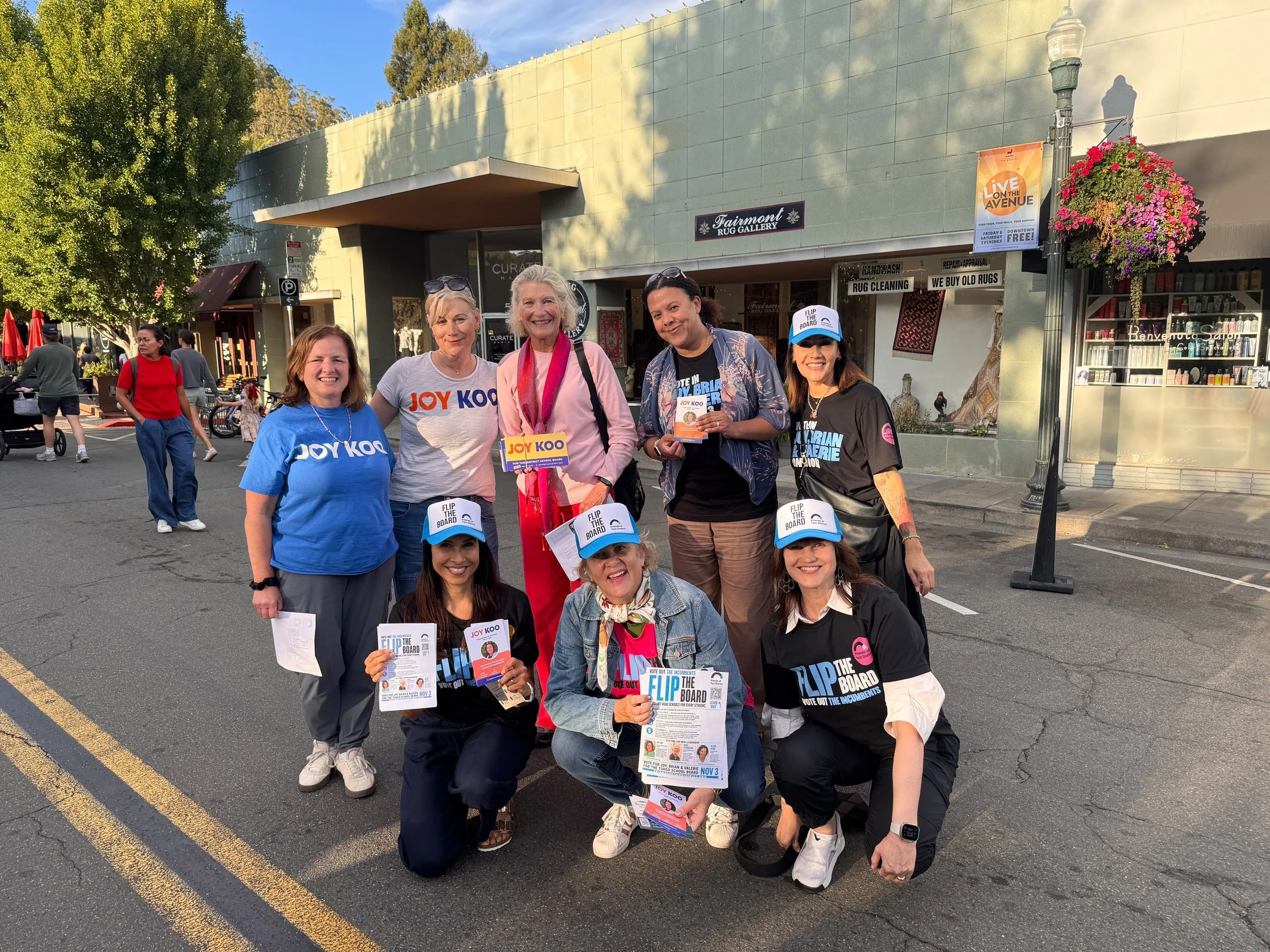 Group of people standing outside a shopping center holding flyers advocating for voting on a school board issue. Five individuals are smiling and posing for the photo, with some wearing blue shirts and campaign signs that say 'Joy Koo' and 'Flip the 