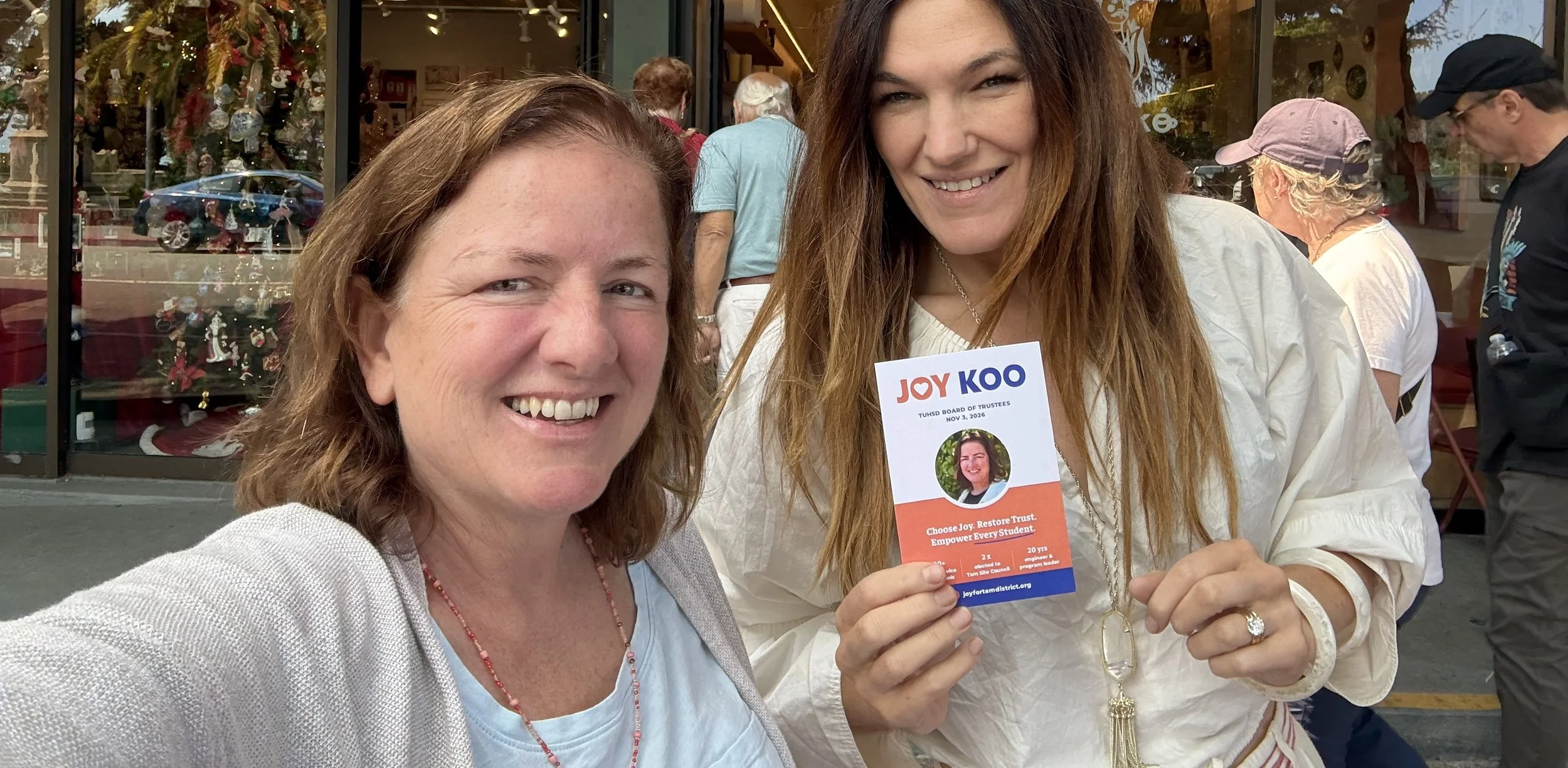 Two women smiling and taking a selfie outdoors. One woman holds a campaign flyer for Joy Koo, showcasing her picture and political message. People are visible in the background, standing in front of a store with holiday decorations.