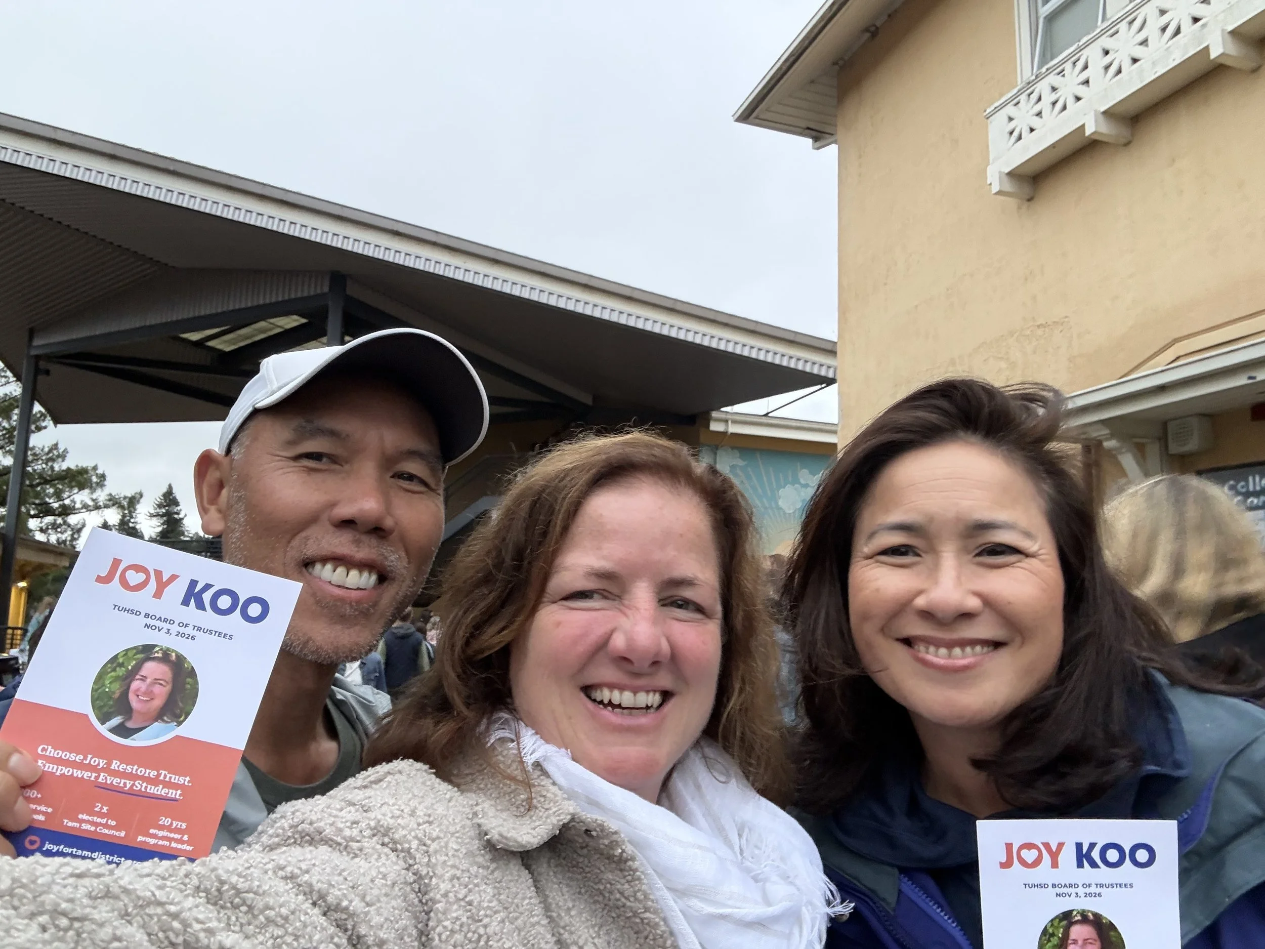 Group of smiling people outdoors holding campaign flyers for Joy Koo, at a public event. Overcast sky with building in background.
