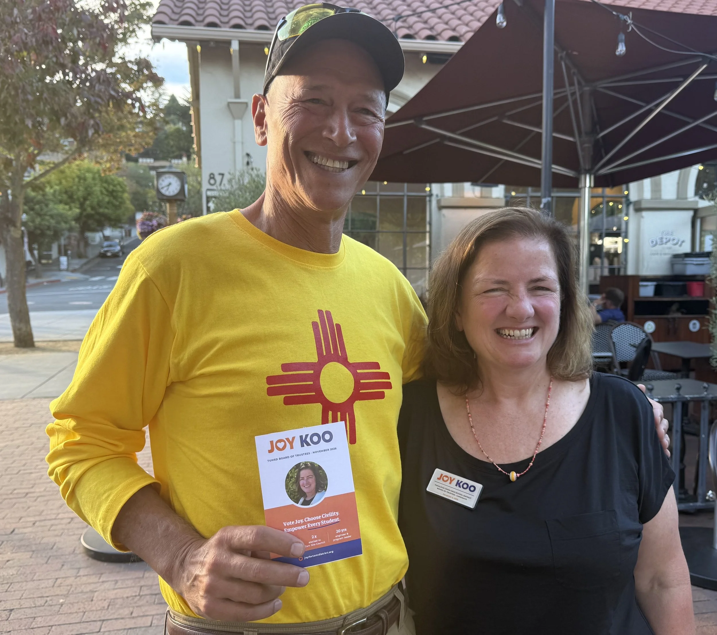 Close-up of an older man in a yellow T-shirt and a baseball cap, smiling and holding a political campaign flyer with his left hand. A woman with brown hair and a black shirt is partly visible next to him, also smiling. They are standing outdoors in a