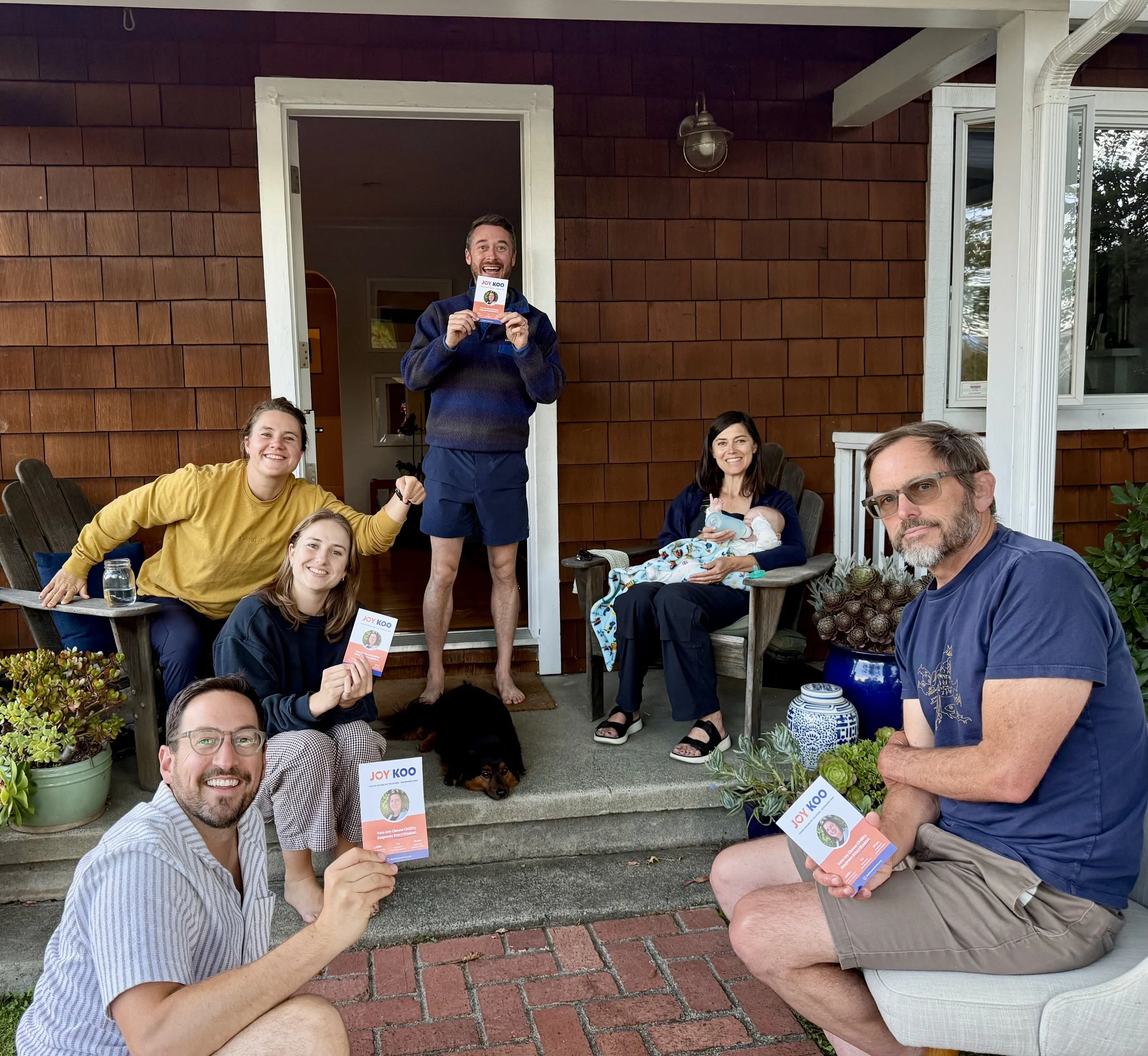 Group of people sitting and standing on a porch with red brick and wooden exterior, holding flyers, smiling, with plants and trees in the background.