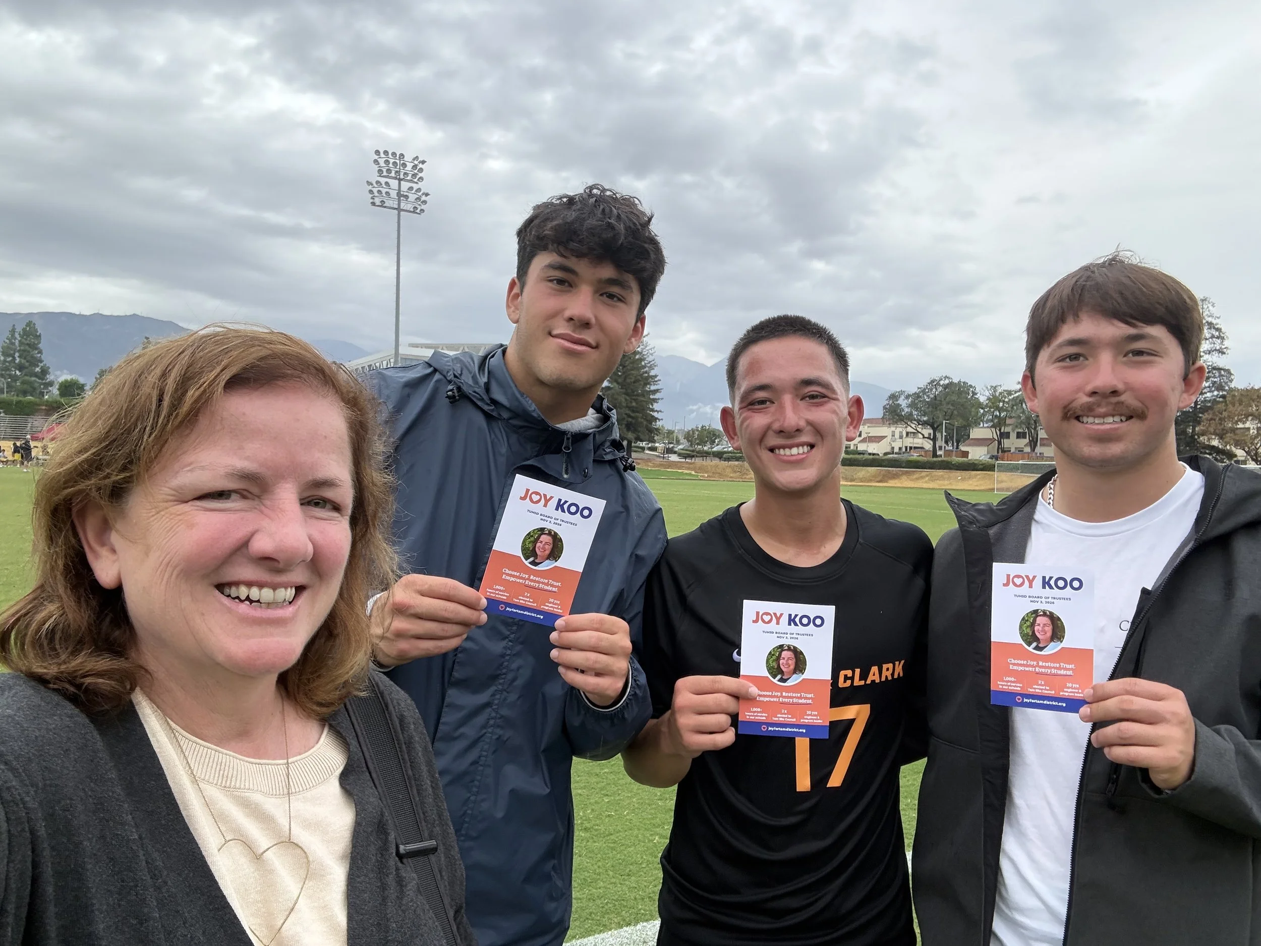 A group of four smiling people outdoors at a sports field on a cloudy day, holding flyers for Joy Koo with a mountain range and stadium lights in the background.