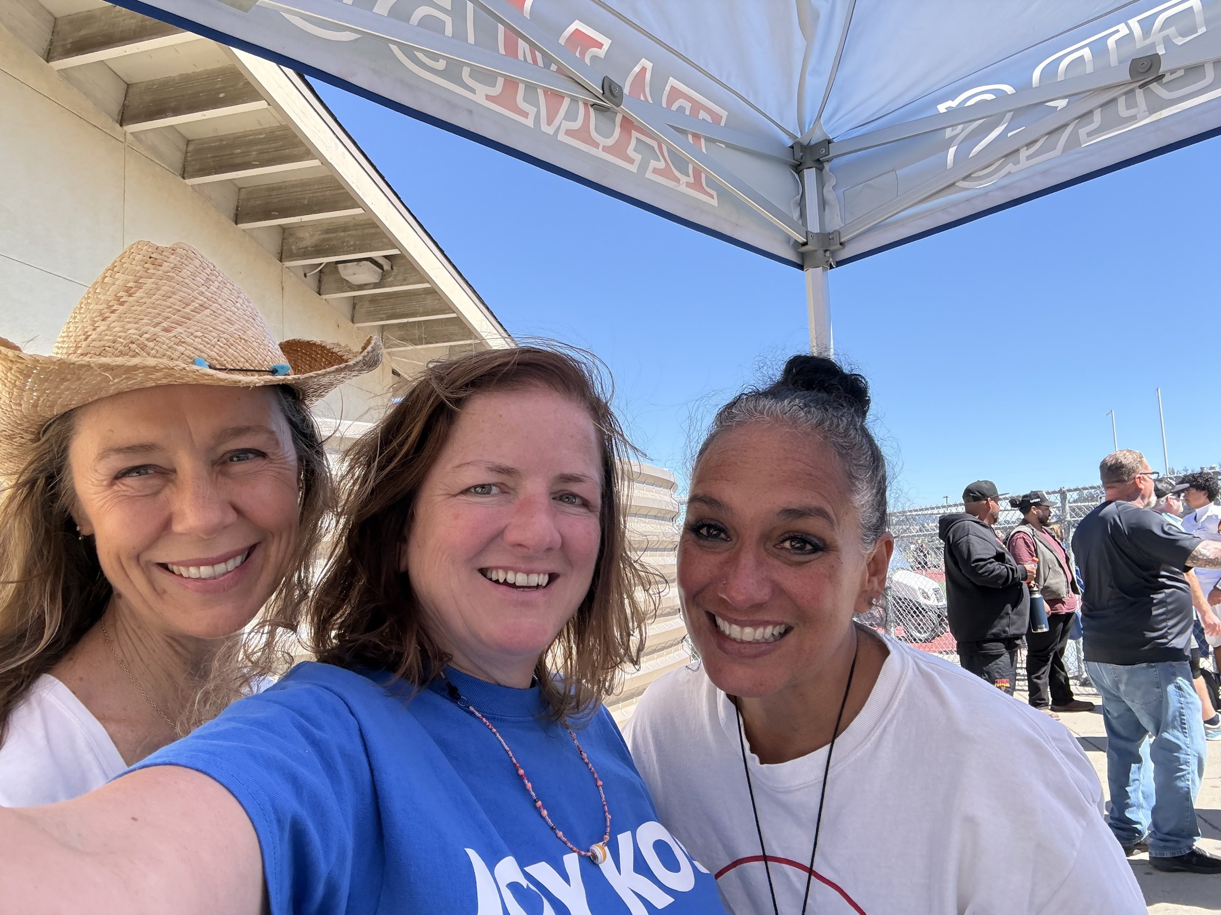 Four women smiling and taking a selfie outdoors at a sunny event. One woman is wearing a straw hat, and another is wearing a blue shirt. There is a crowd of people and a chain-link fence in the background with a clear blue sky above.