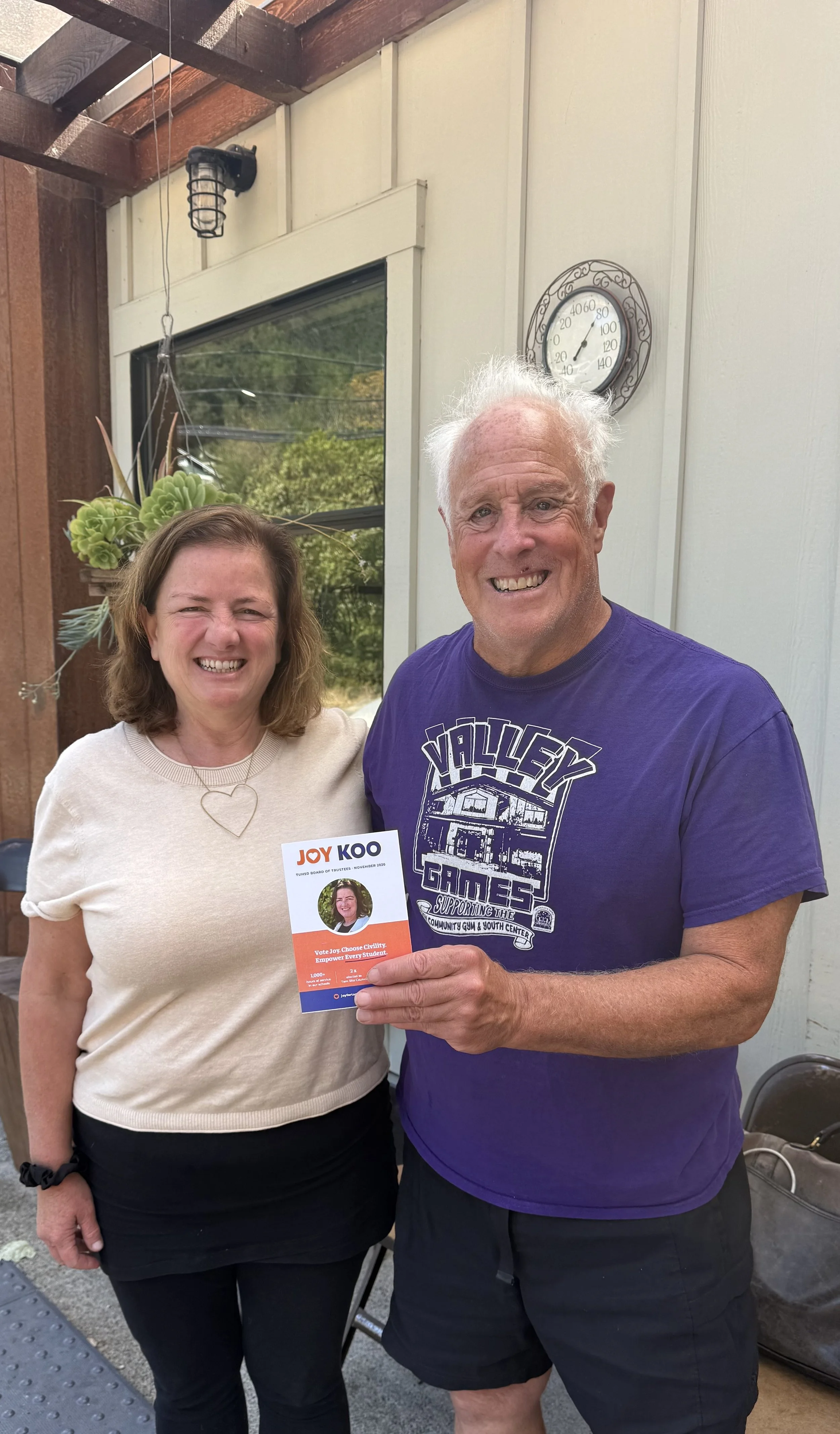 A woman and a man are smiling and posing for a picture outdoors. The woman is holding a voter registration card, and the man is pointing at it. The woman has shoulder-length brown hair and is wearing a beige sweater and black pants. The man has white