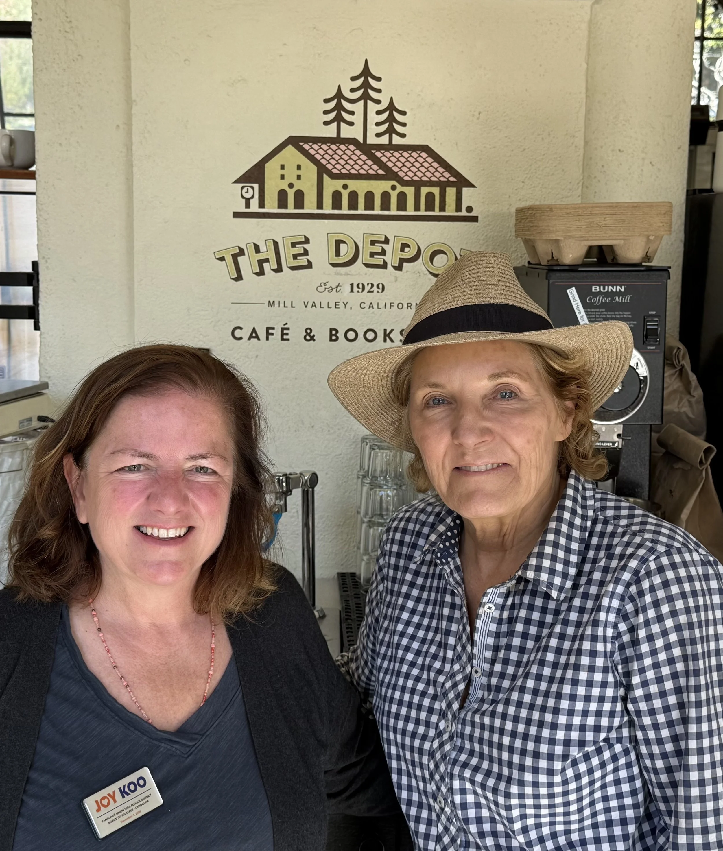 Two women smiling for a selfie at a cafe with a sign reading 'The Depot' in the background, one wearing a straw hat and checkered shirt, and the other wearing a name tag that says Joy Koo.