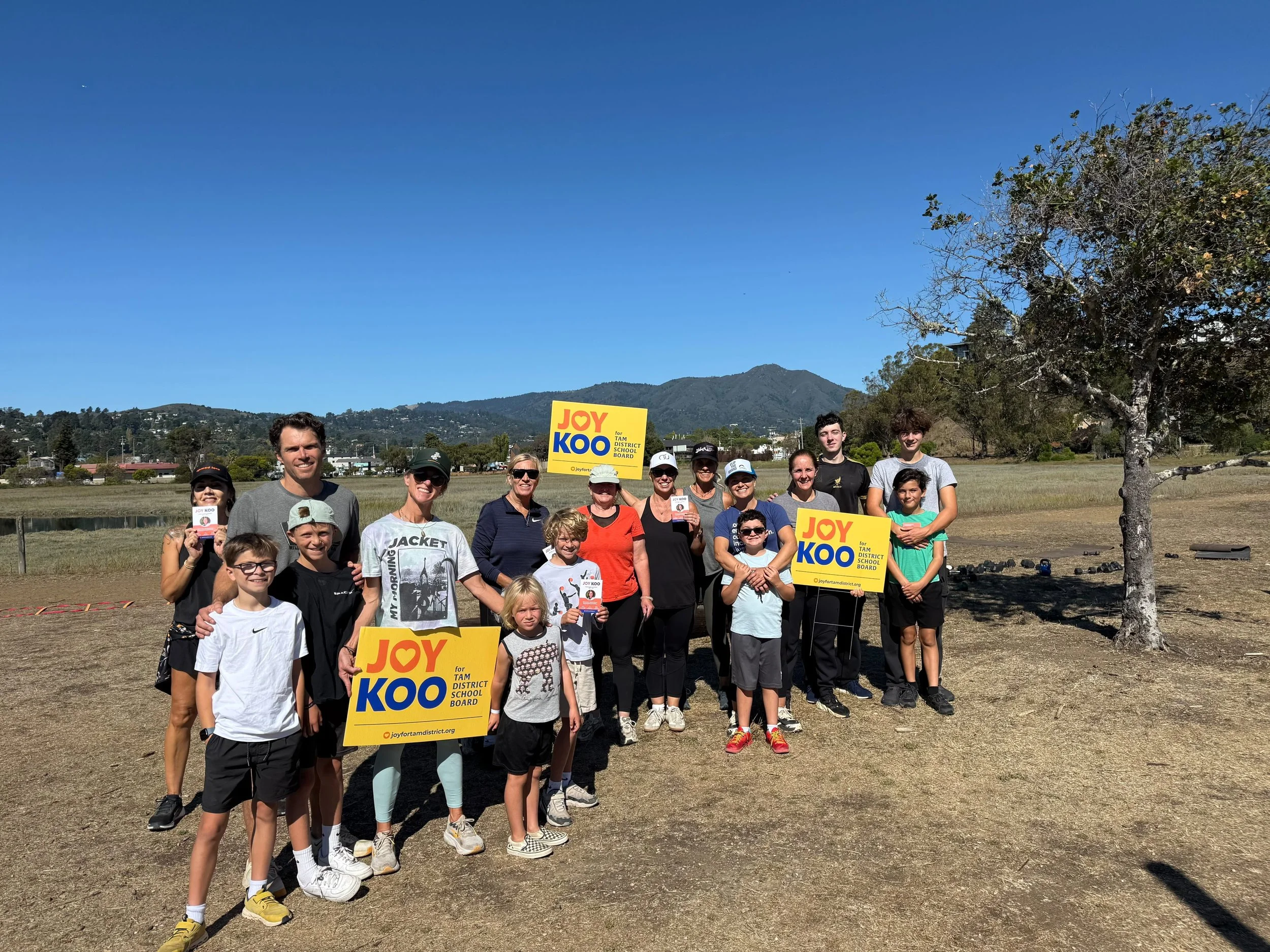 A group of people gathered outdoors on a sunny day with mountains and trees in the background. Some are holding signs that say 'Joy Koo for TAM District School Board'.