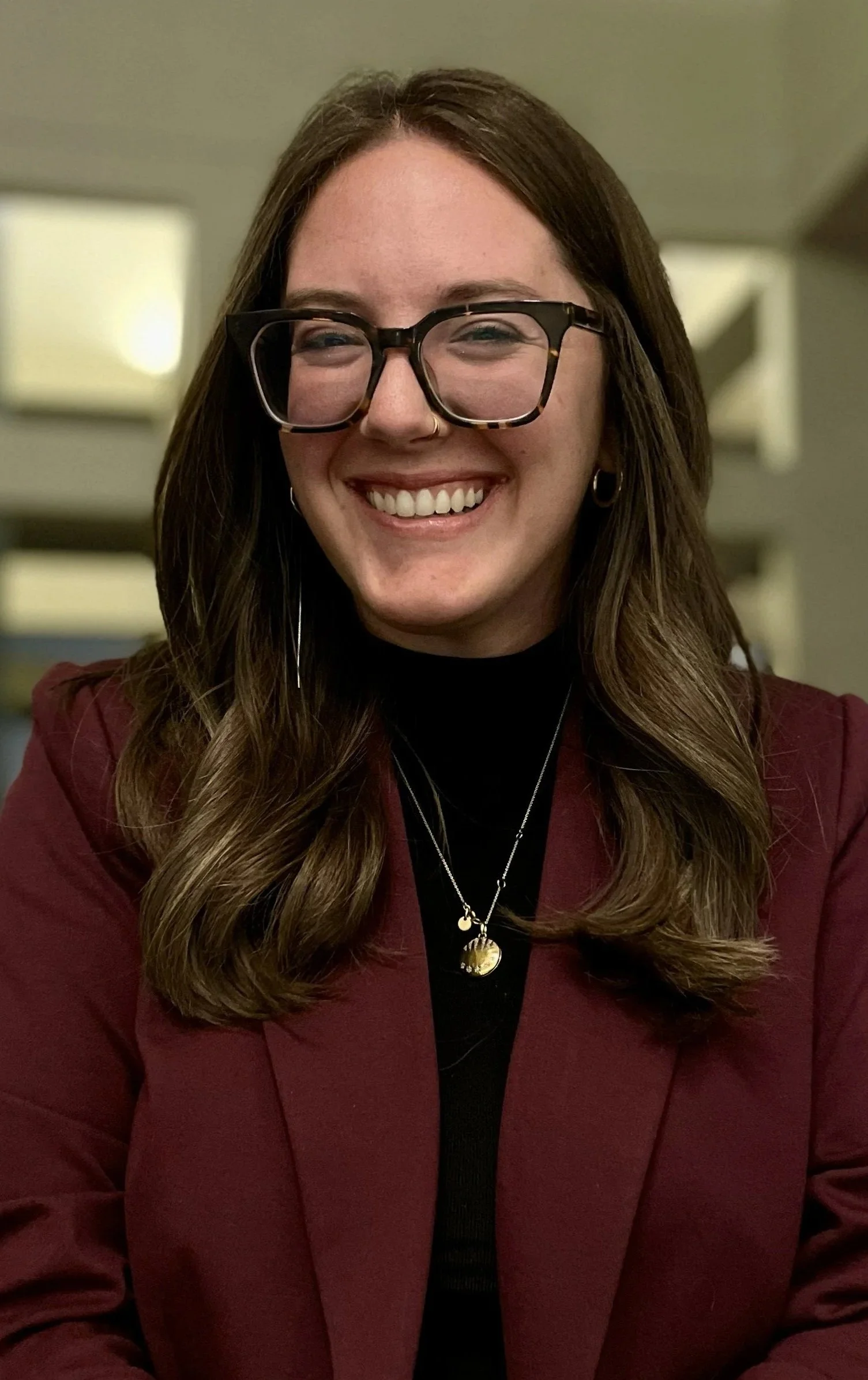 A smiling woman with long wavy brown hair, wearing large tortoise-shell glasses, a nose ring, hoop earrings, a black turtleneck, a burgundy blazer, and layered necklaces, indoors with blurred background.