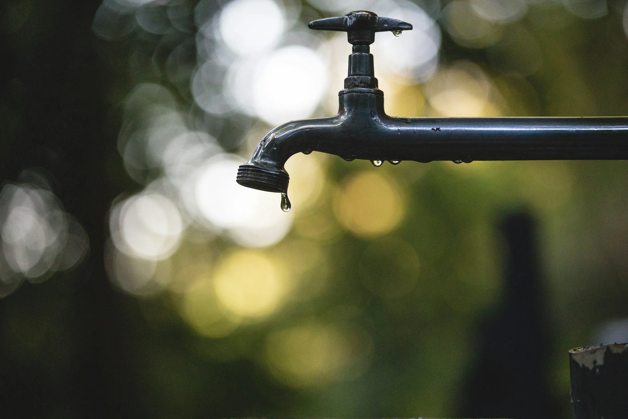 Close-up of a black outdoor water faucet with water dripping from its spout, against a blurred background of green and white bokeh lights.