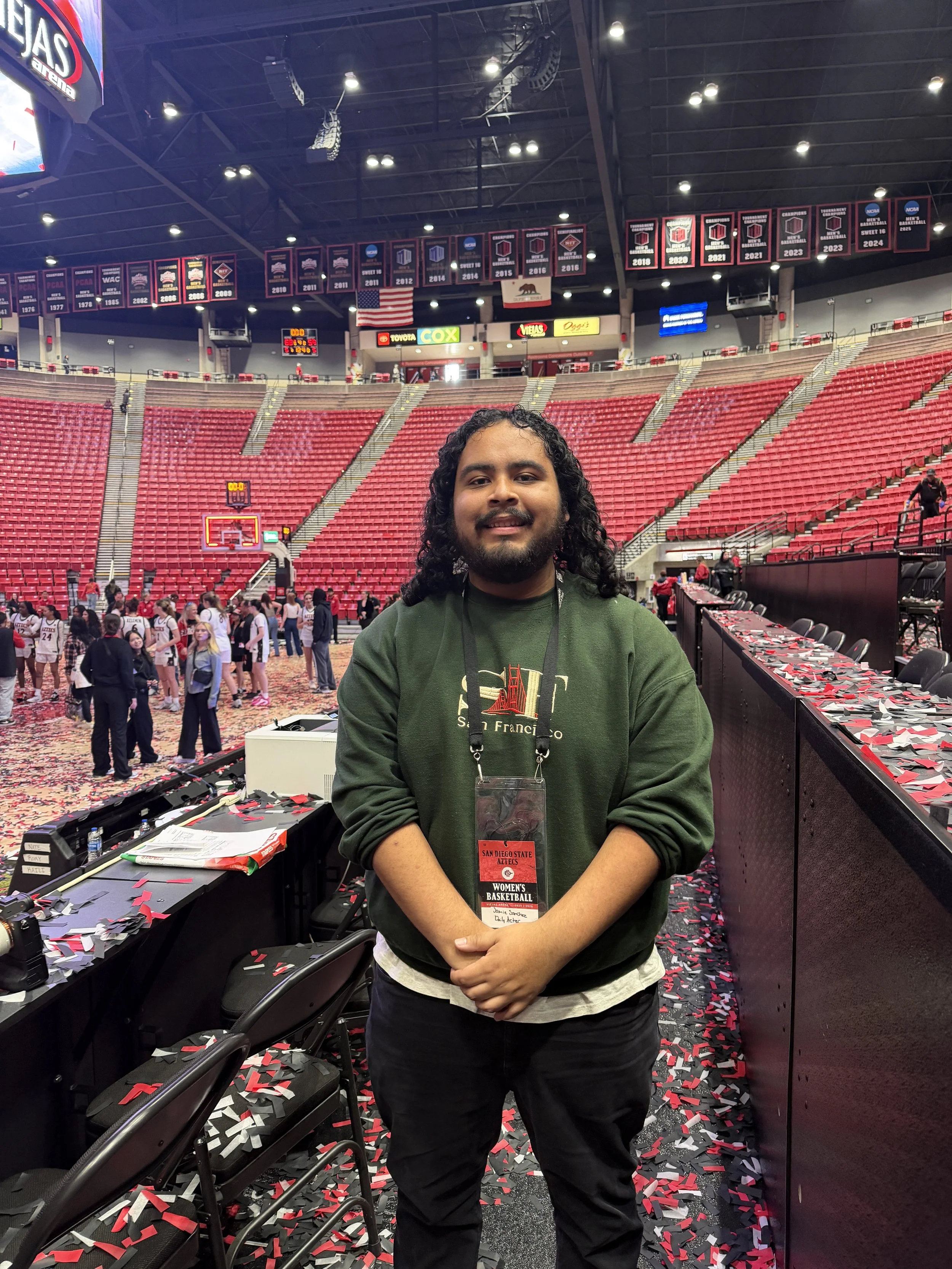 A man with long curly hair and a beard, wearing a dark green sweatshirt and black pants, standing inside a sports arena with red seats and time clock, confetti on the floor, and banners hanging from the ceiling, holding a badge for San Diego State women's basketball.