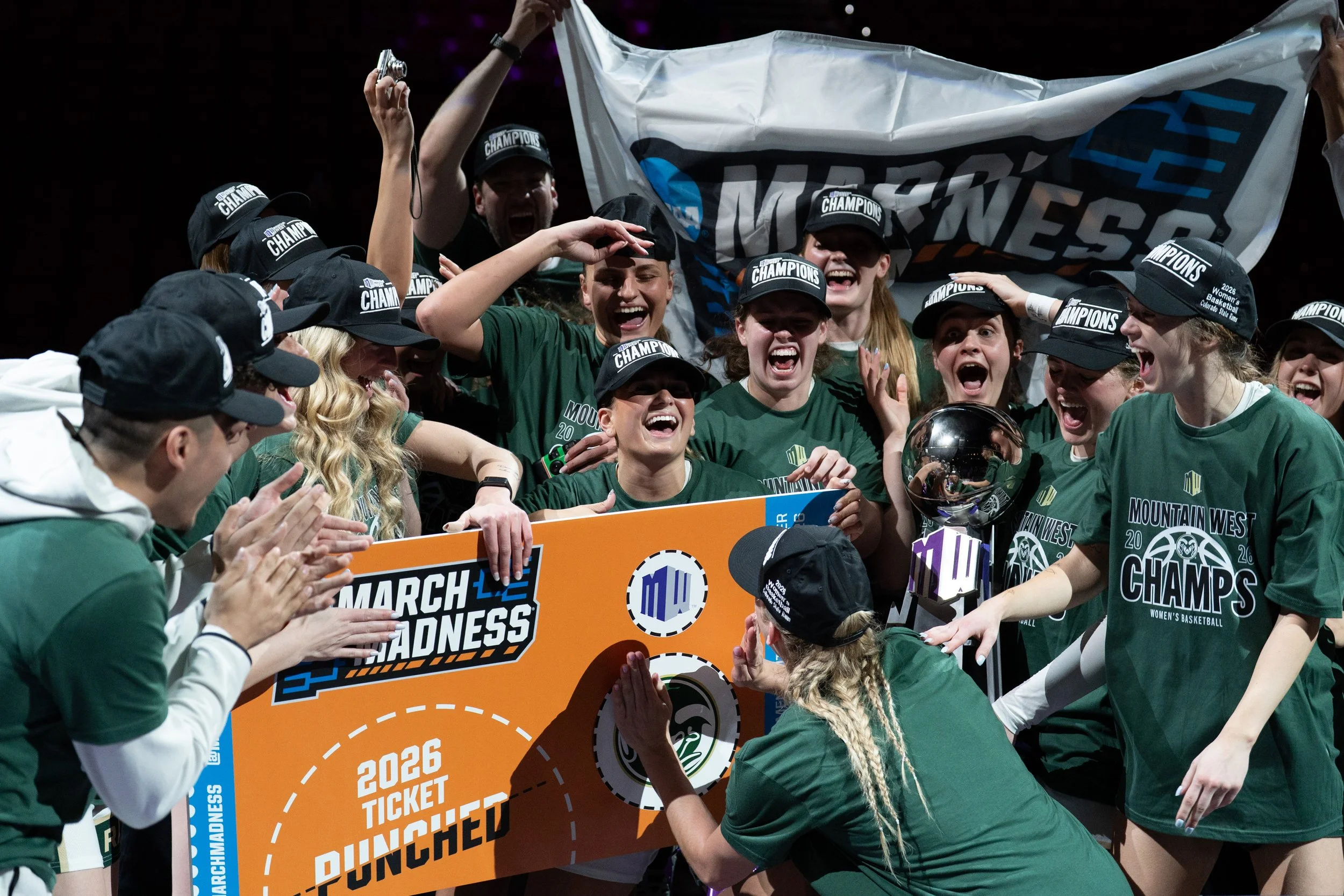 Group of young women and men celebrating victory at a sports championship, wearing green shirts and caps labeled 'Champions,' holding a large orange sign that reads '2026 Ticket Puncher,' with some players reaching out toward each other and a flag in