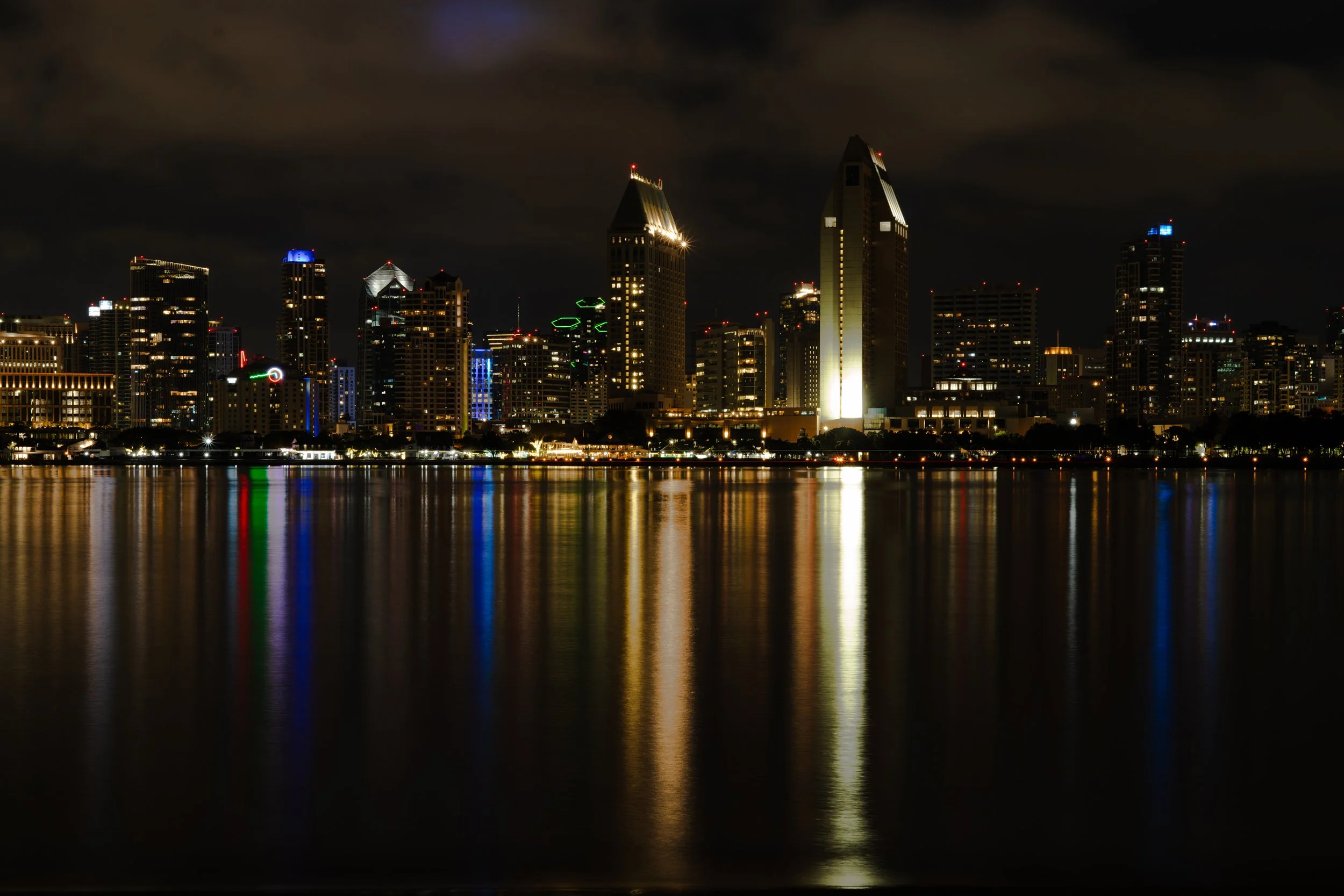 Nighttime city skyline with tall buildings illuminated, reflecting on calm water.