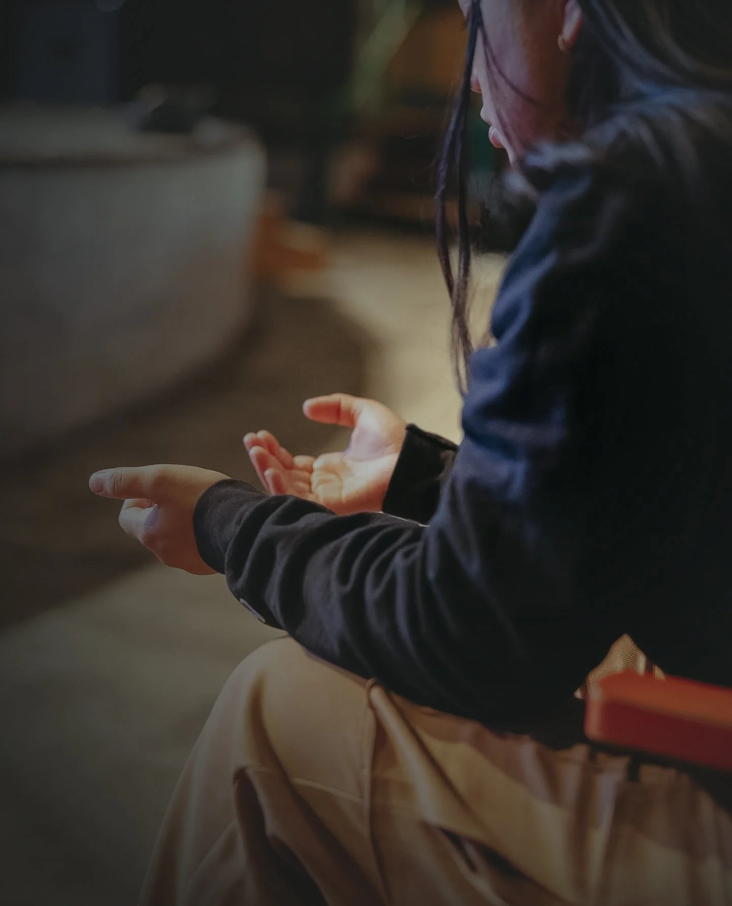 A person with dark hair and glasses, dressed in a dark jacket, sitting on a bench with their hands clasped together as if in prayer or reflection.