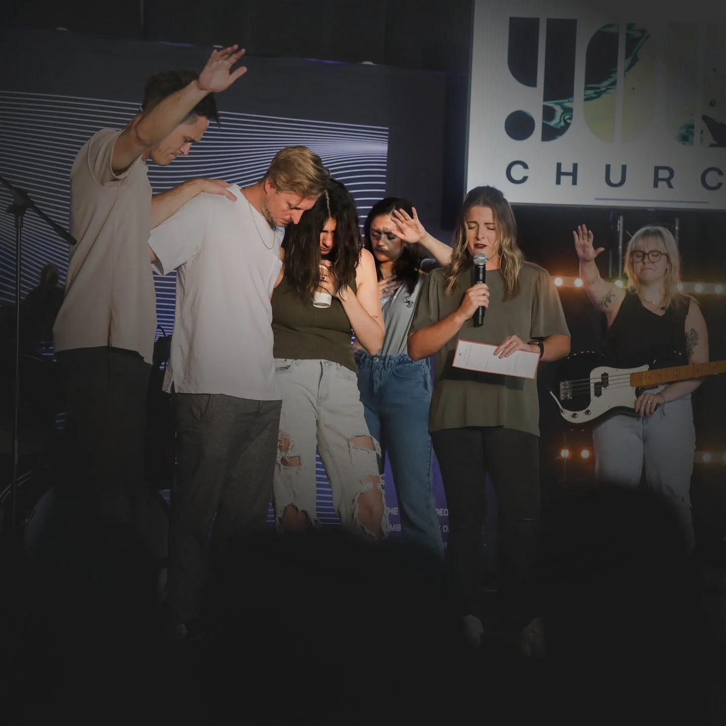 Group of six people with eyes closed or in prayer on stage, some with raised hands, some with closed eyes, a woman with a microphone and script, and a woman holding a bass guitar, in front of a church logo.