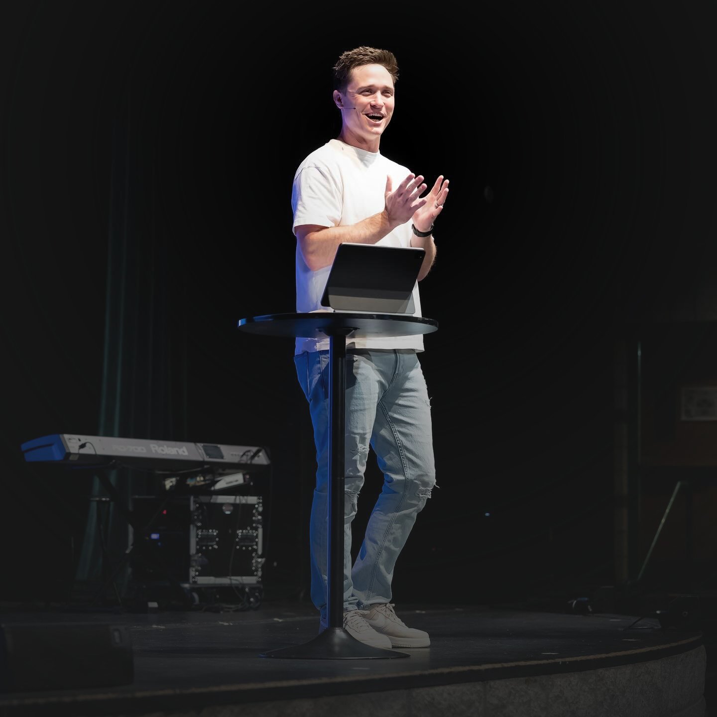 A man standing on stage at a lectern with a tablet, speaking and gesturing with his hands, with musical equipment in the background.
