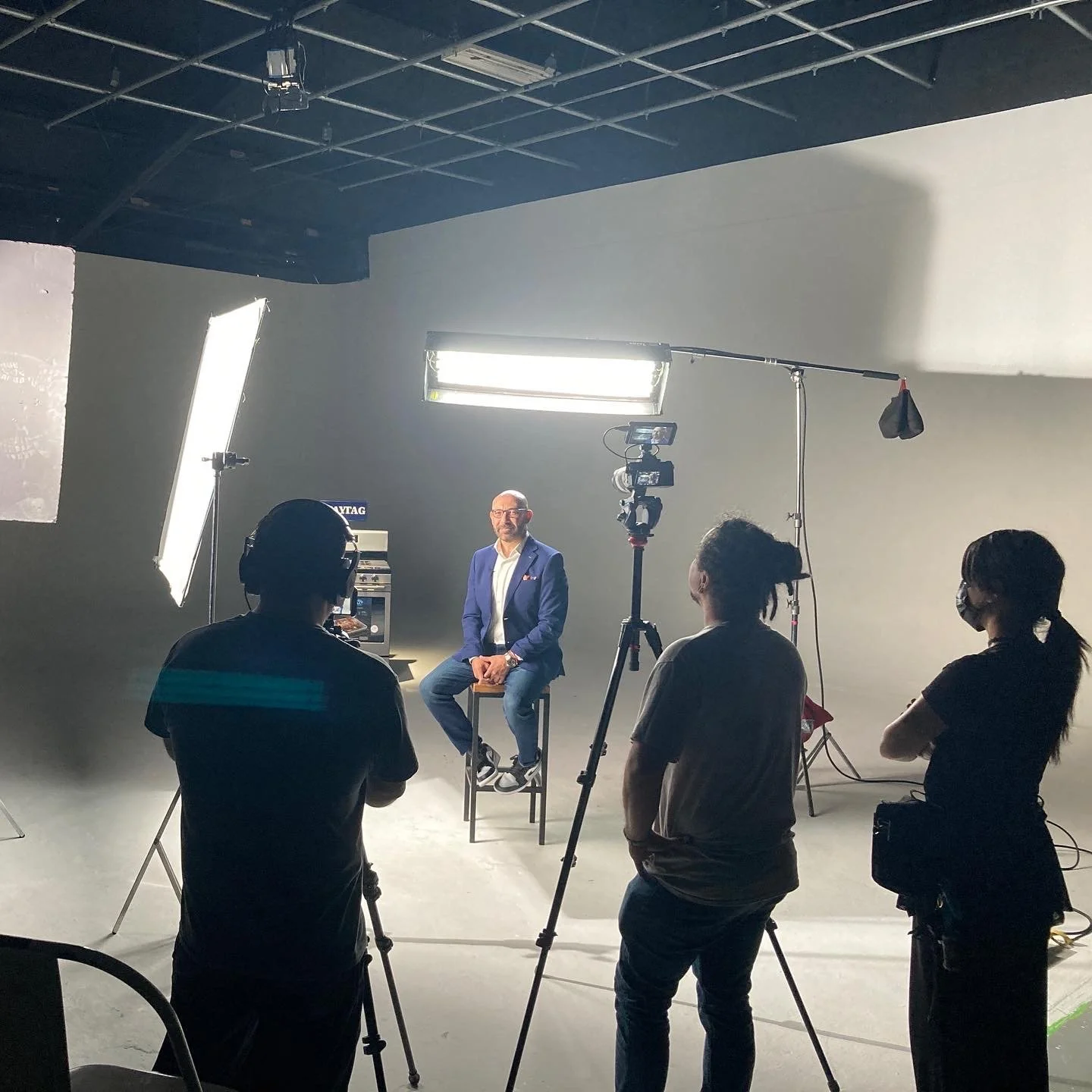 A man in a blue blazer and glasses is seated on a stool in front of a camera, being filmed by a crew of four people in a photography studio with professional lighting.
