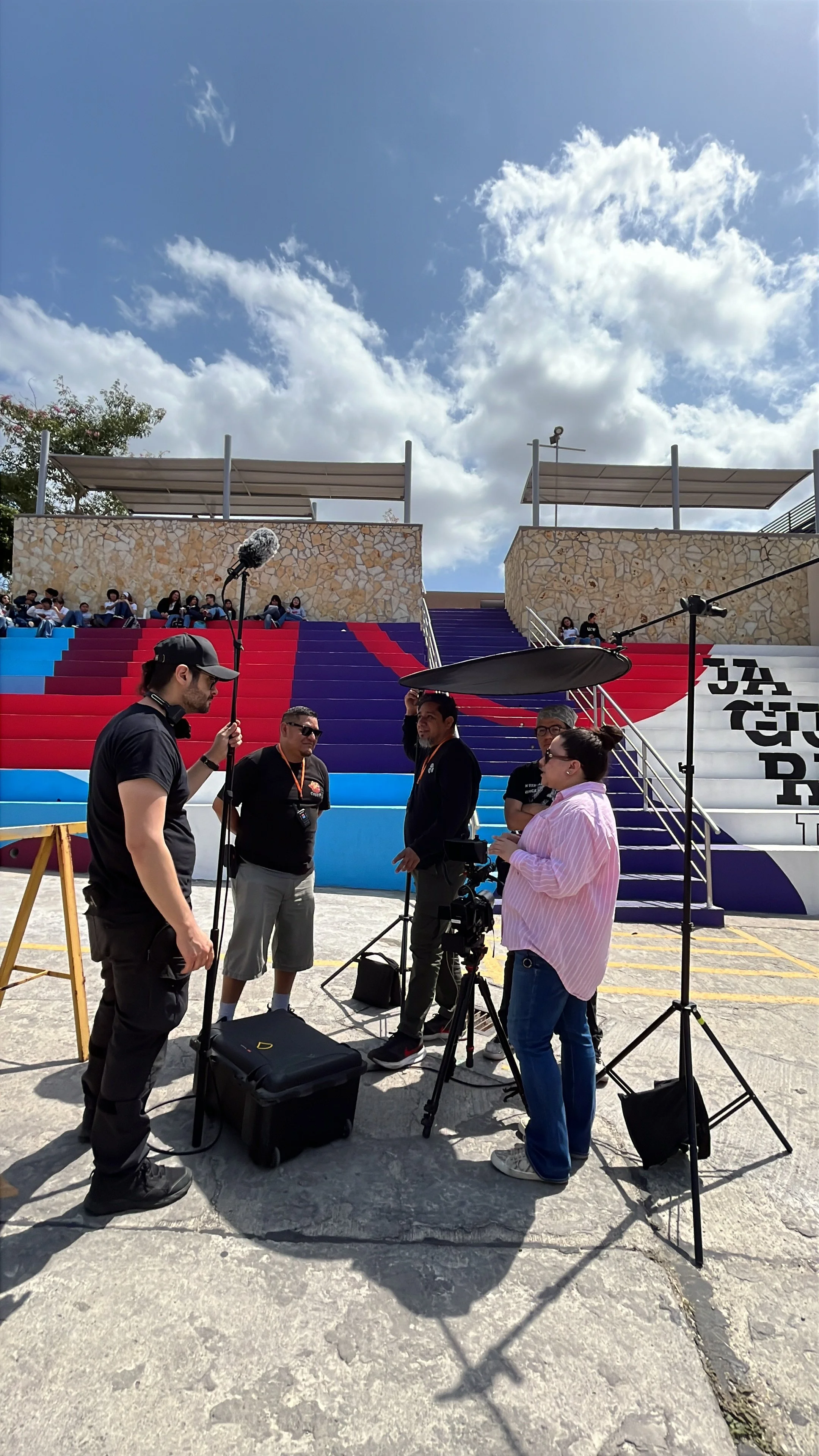 A group of people filming a video or conducting an interview outdoors. There are multiple cameras, lighting equipment, and crew members gathered around a colorful stairway with red, purple, and blue steps, and a wall with text on the right side.