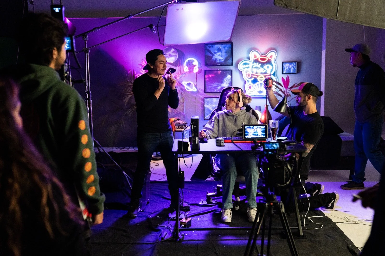 Filming set with crew and actors, purple neon lights, and a woman with headphones at a desk.