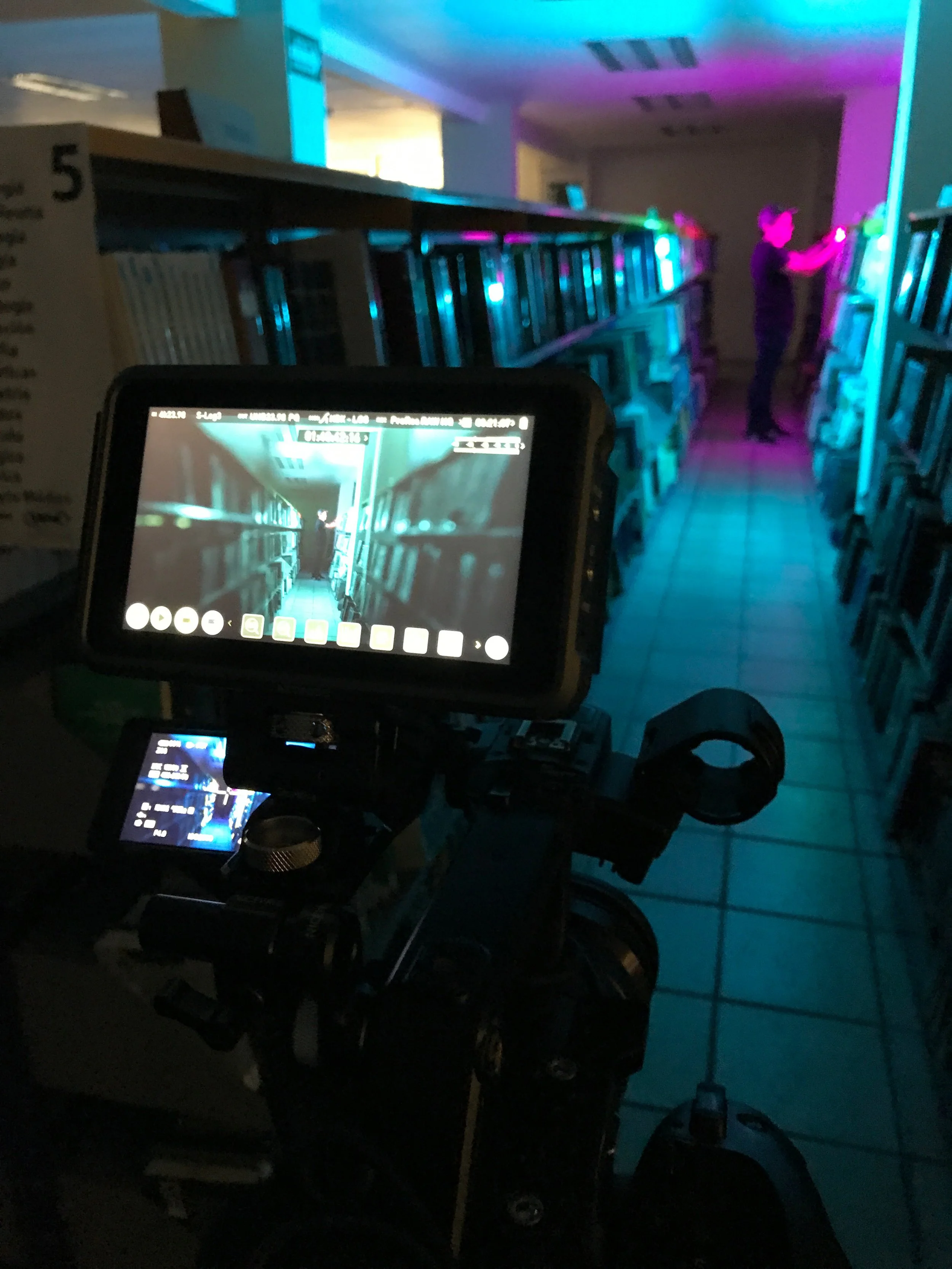 Photo taken in a dimly lit room with colorful LED strip lights, showing a person in dark clothing using a vending machine or similar equipment, with a monitor displaying a digital interface in the foreground.