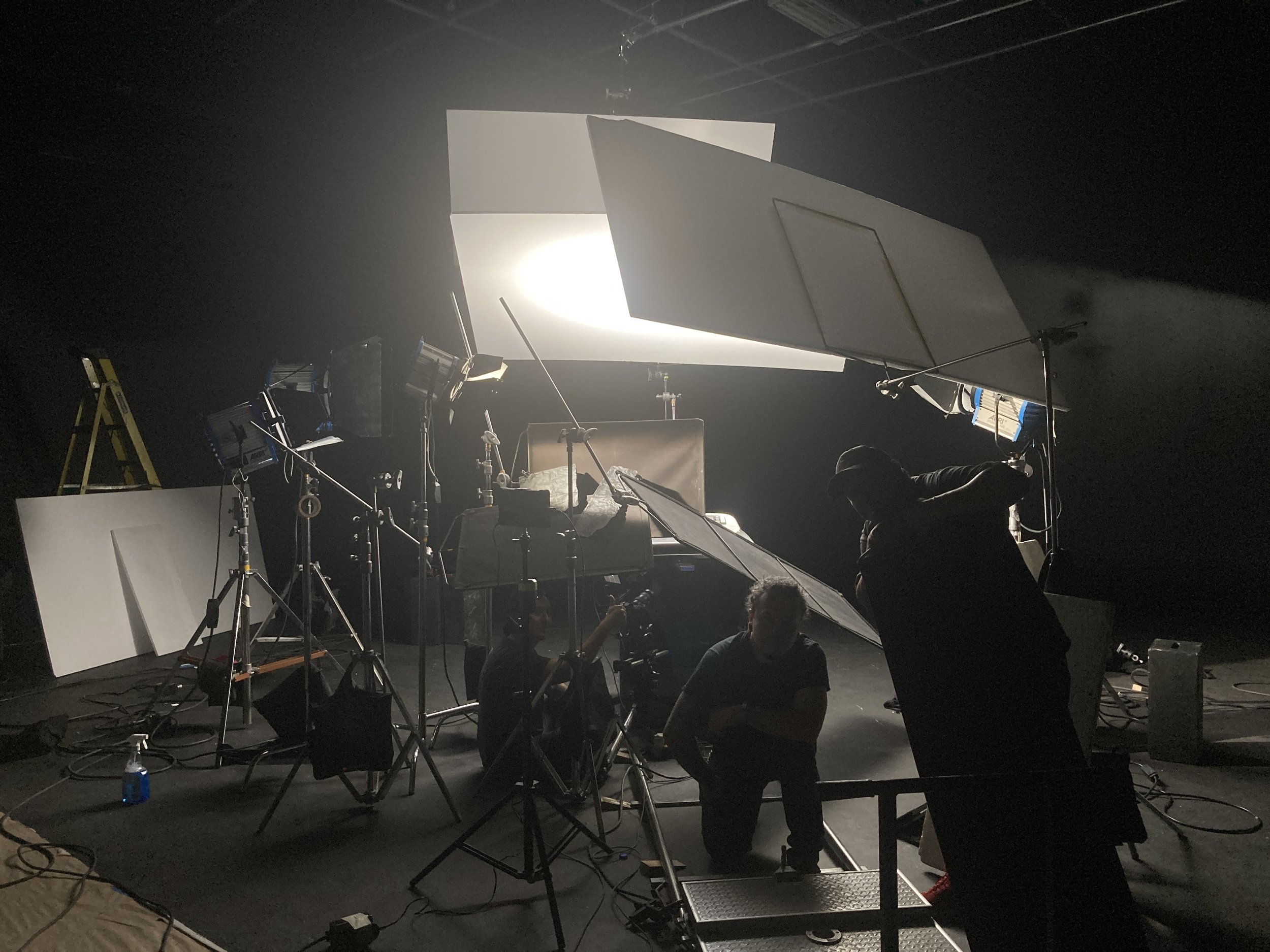 View of a photography or film studio with large studio lights, reflectors, and backdrop. Several crew members are preparing the setup, with equipment and cables visible on the floor.