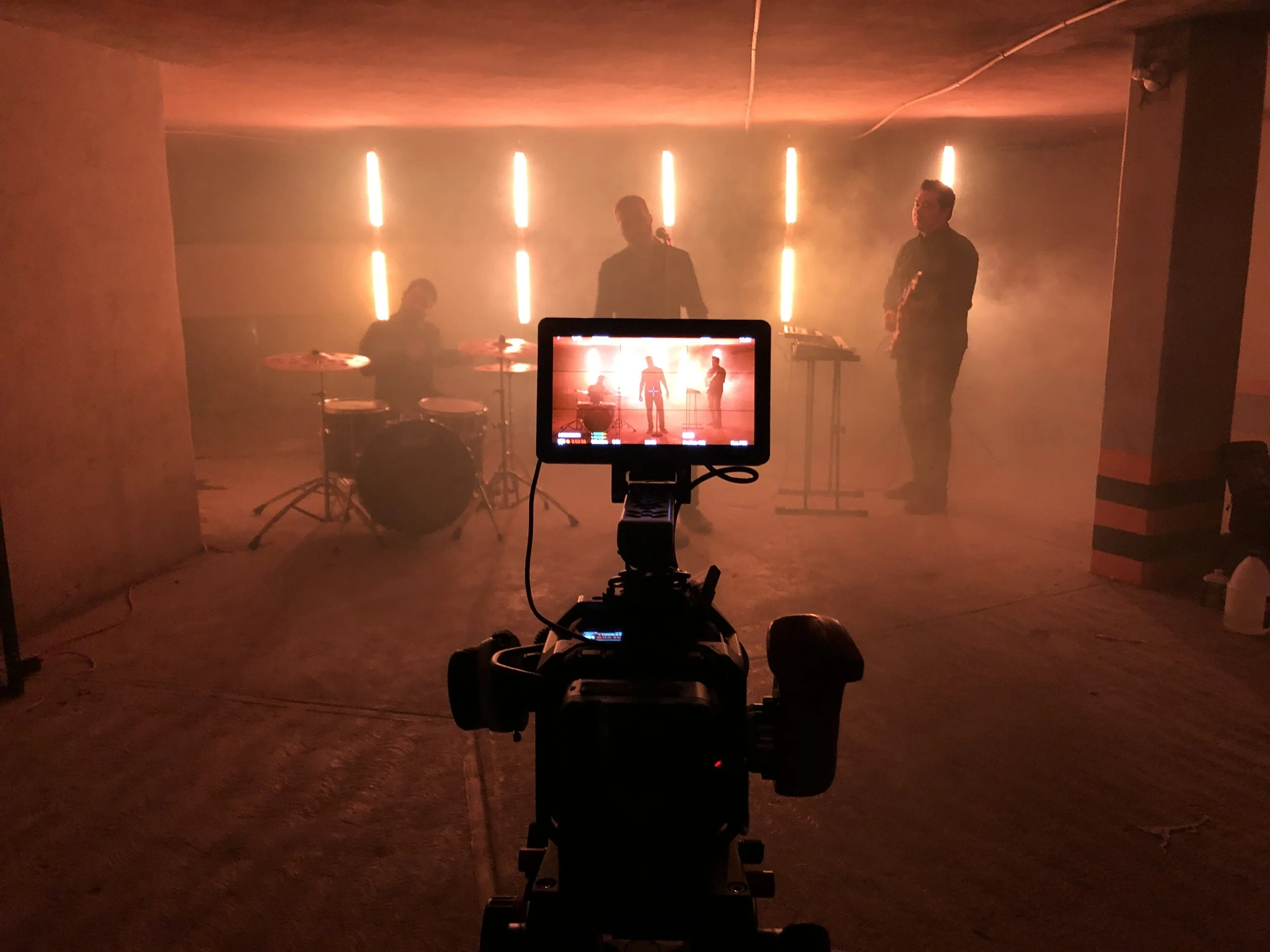 A music band performing on stage with three musicians, one playing drums, one singing into a microphone, and another standing with a keyboard, illuminated by vertical orange light bars, with a camera recording from in front.