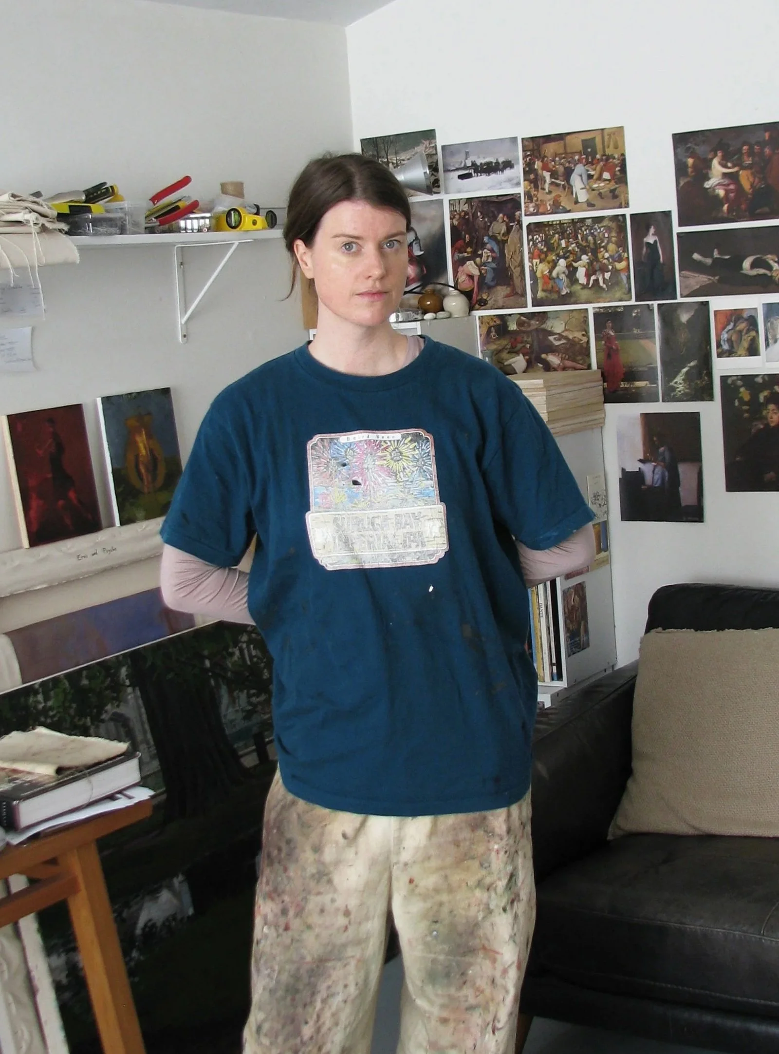Chloe Summerhayes in a studio, wearing a blue t-shirt over a long sleeve shirt and paint-splattered pants.
