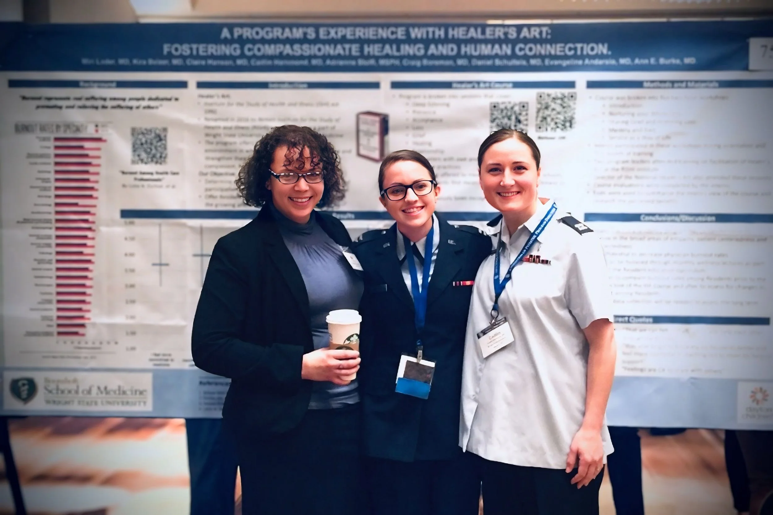 Three women standing in front of a large informational poster about healer's art and compassionate healing, smiling at the camera. Dr. Lader, to the left, is holding a cup of coffee.