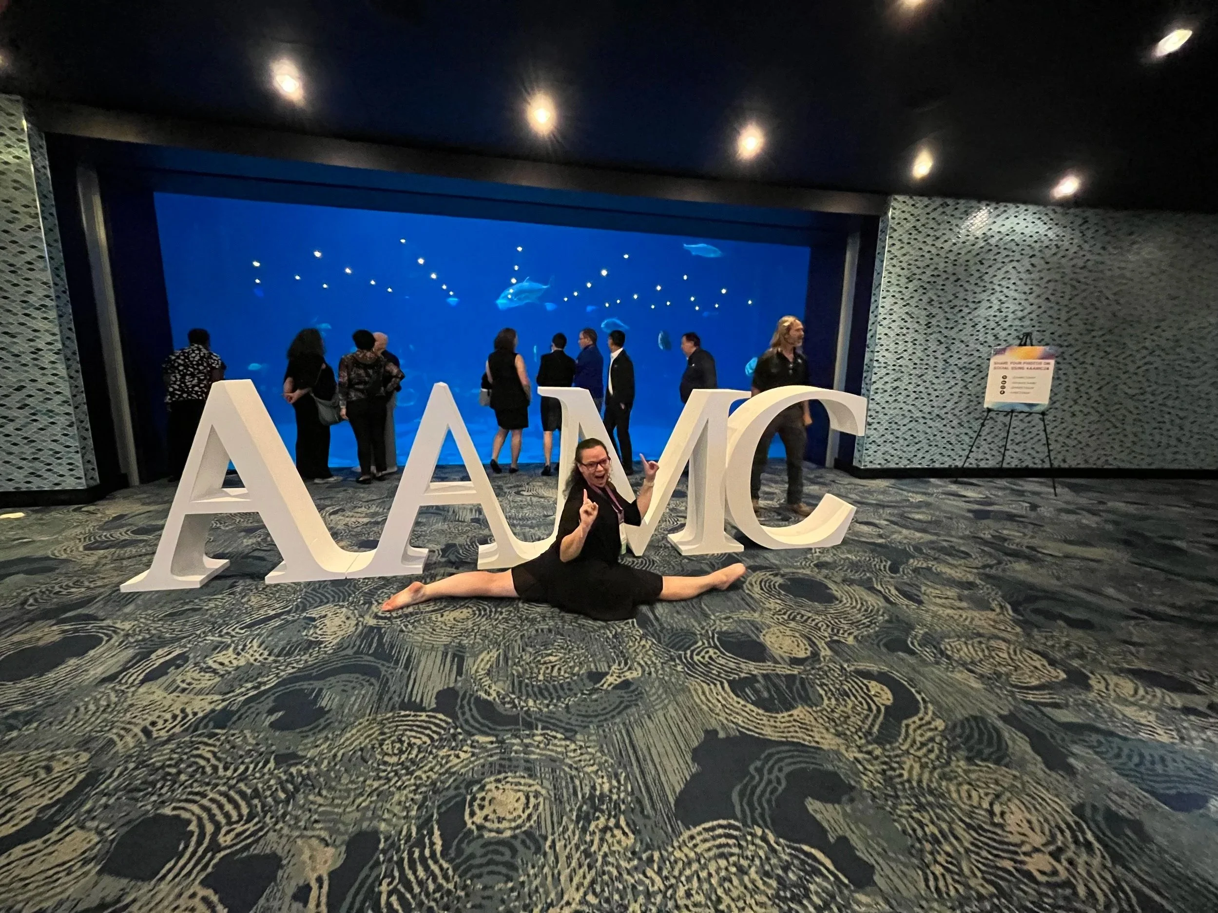 Dr. Lader performs a split on the carpeted floor of a conference, in front of large white letters spelling 'AAMC'. Behind her, people are observing an aquarium tank with fish and blue water.