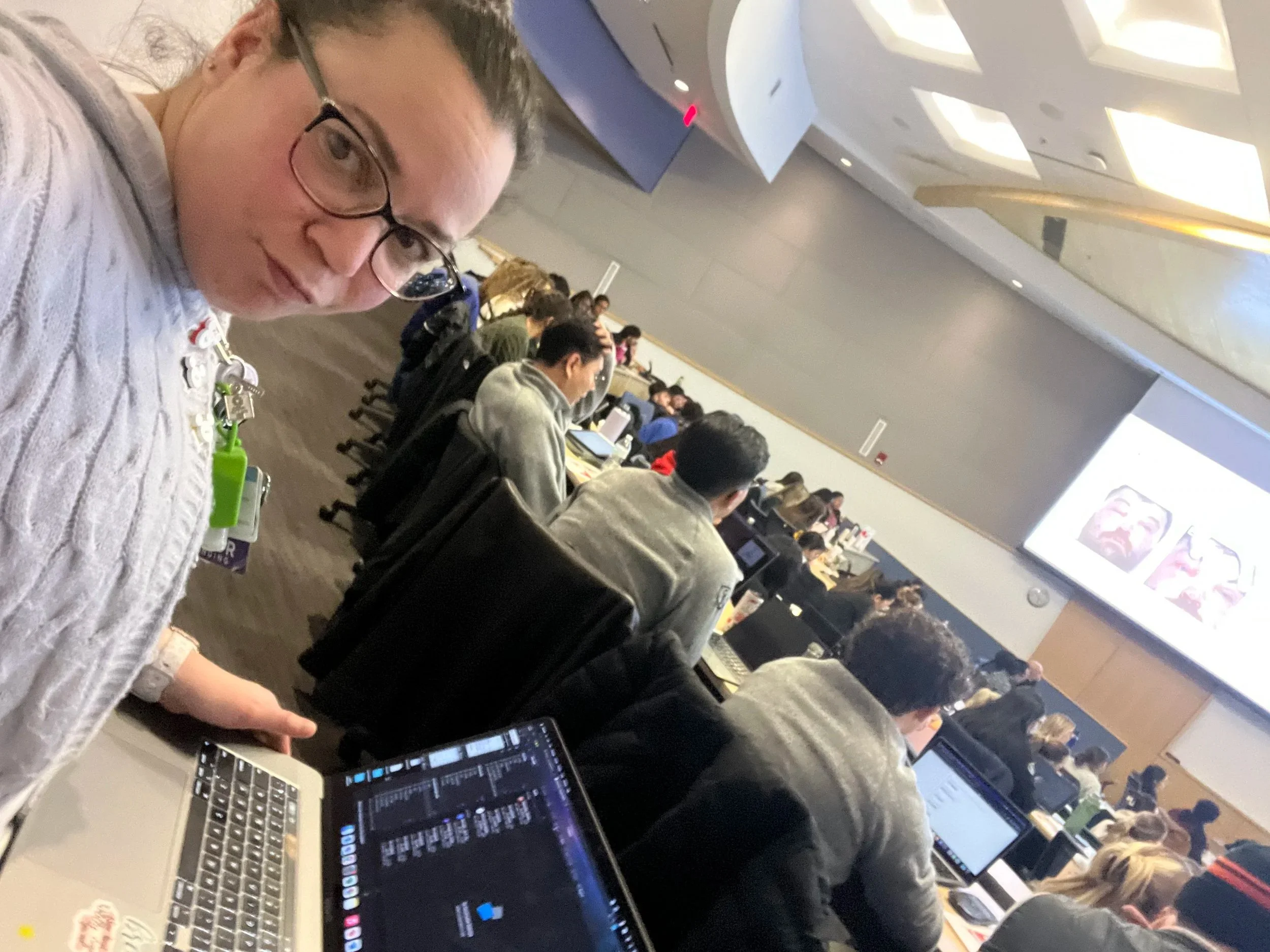 A woman with glasses and curly hair taking a selfie at a conference or workshop. She is sitting at a table with a laptop, and there are other people working on laptops in the background. The room has high ceilings with skylights and a large projection screen showing colorful graphics.