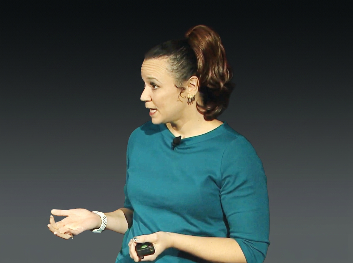 A woman with dark hair tied in a high ponytail, wearing a teal long-sleeve shirt, speaking and gesturing with her hands against a dark background.
