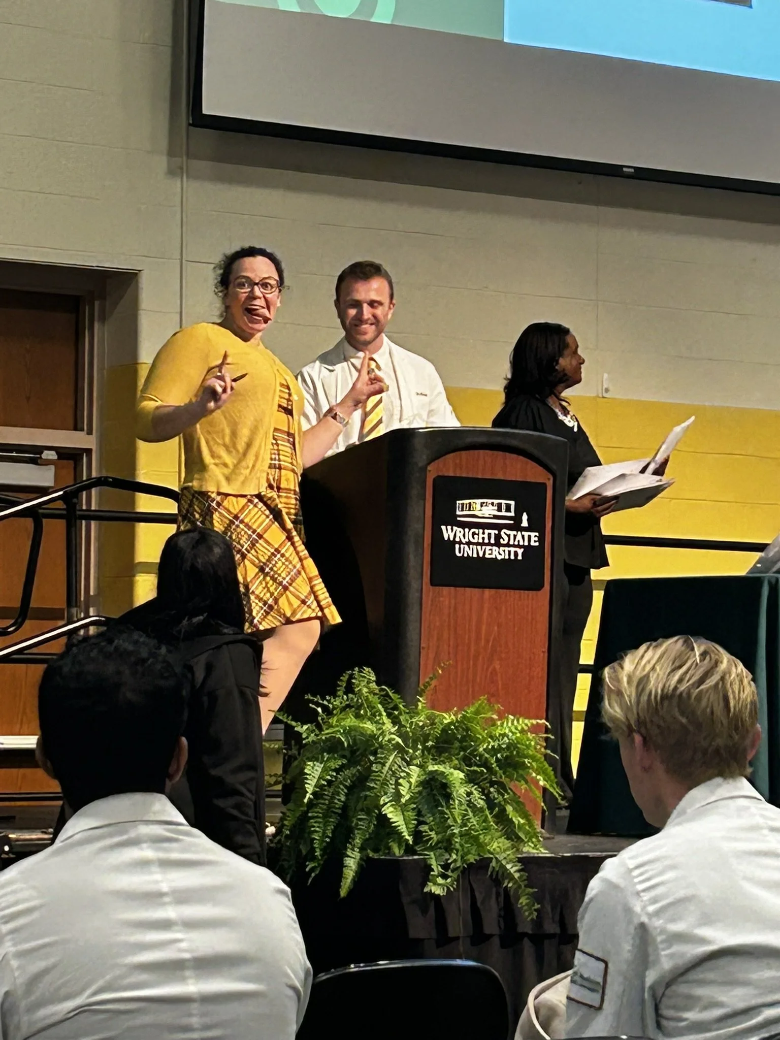 People standing at a podium during an event at Wright State University, with an audience seated in front, and a large screen behind displaying colored graphics.