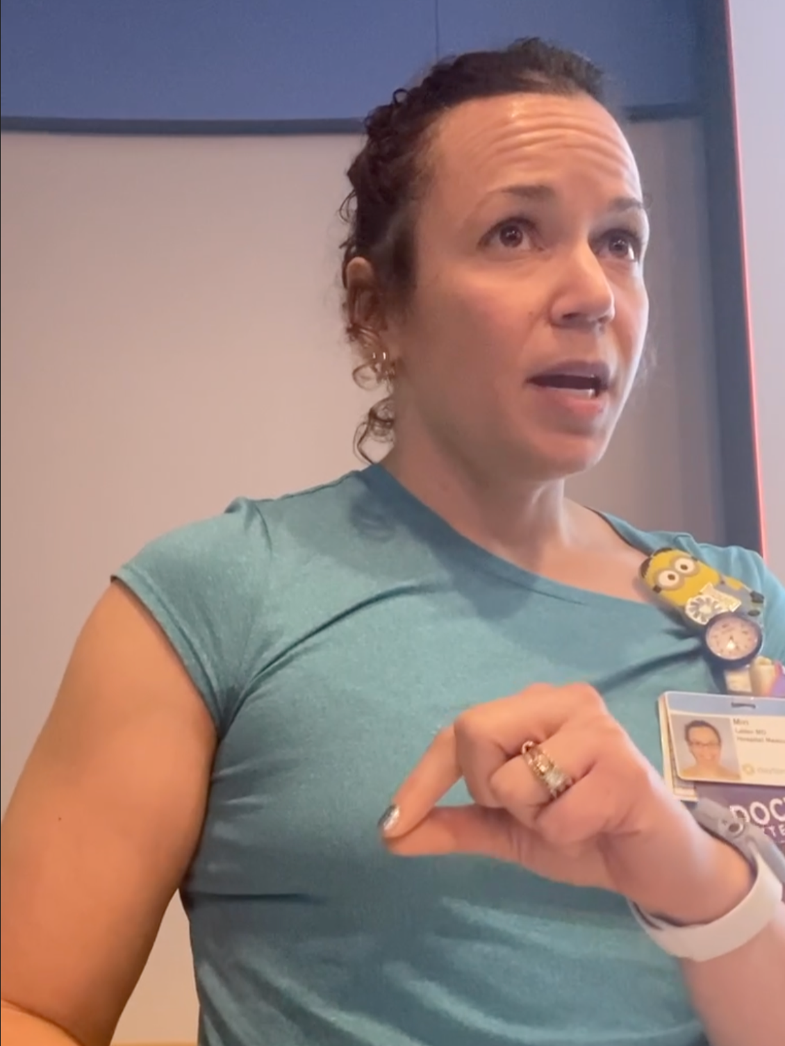Dr. Lader, with curly hair wearing a teal shirt with an ID badge, speaking at a podium in a professional setting.