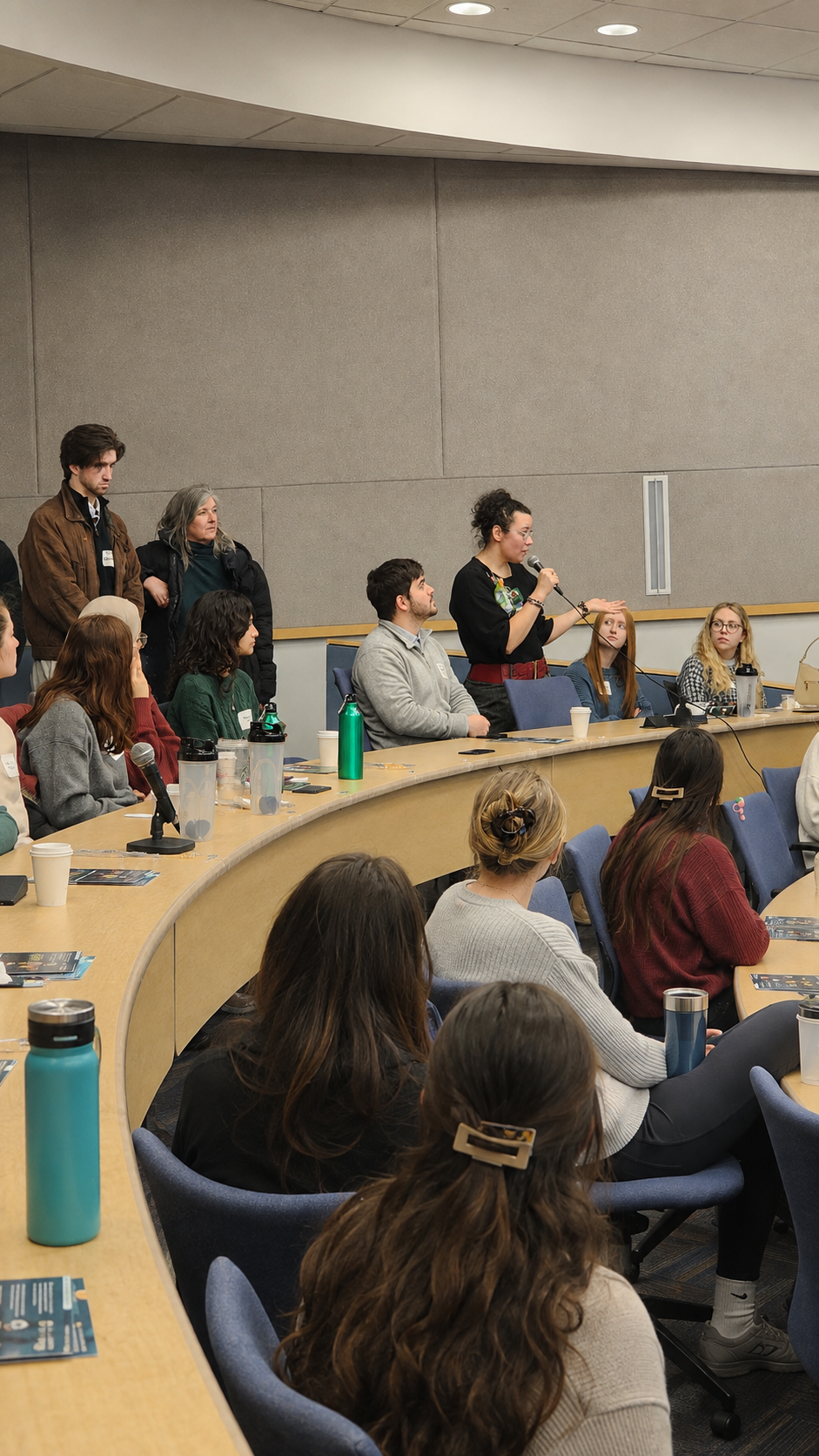 A woman standing and speaking into a microphone at a panel discussion or conference, with seated attendees listening attentively, some taking notes or with drinks on the table.