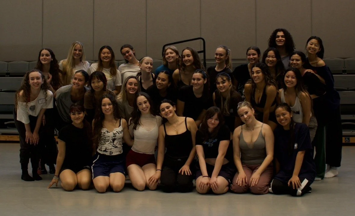 Group of young women and girls posing on a stage in a gymnasium or auditorium.