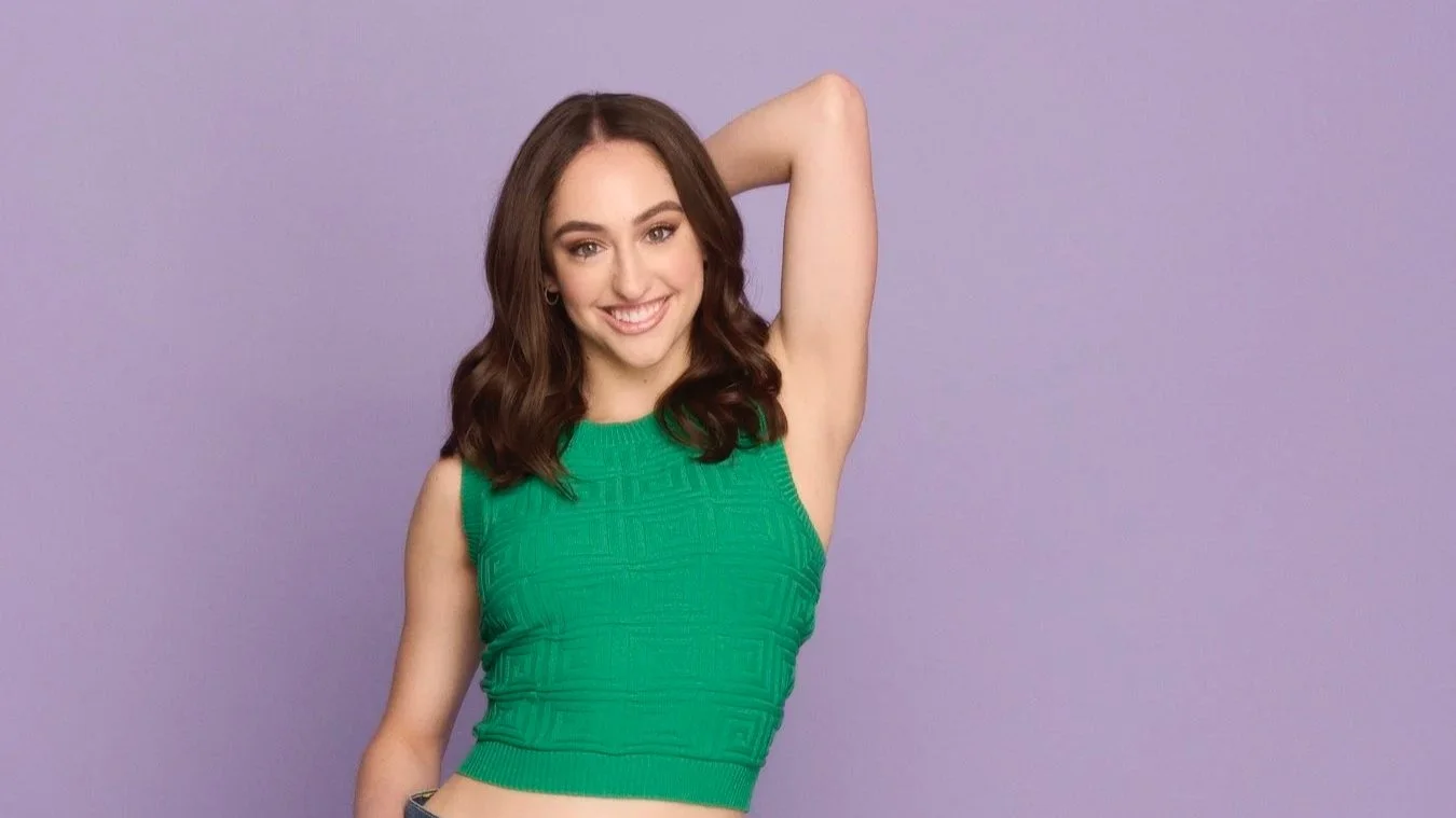 A young woman with brown hair wearing a sleeveless green top, smiling with one arm raised and hands behind her head, standing against a purple background.