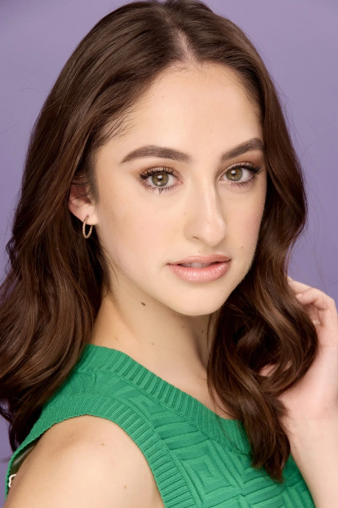 A young woman with long, wavy brown hair and hazel eyes, wearing a green sleeveless top and hoop earrings, posing against a purple background.