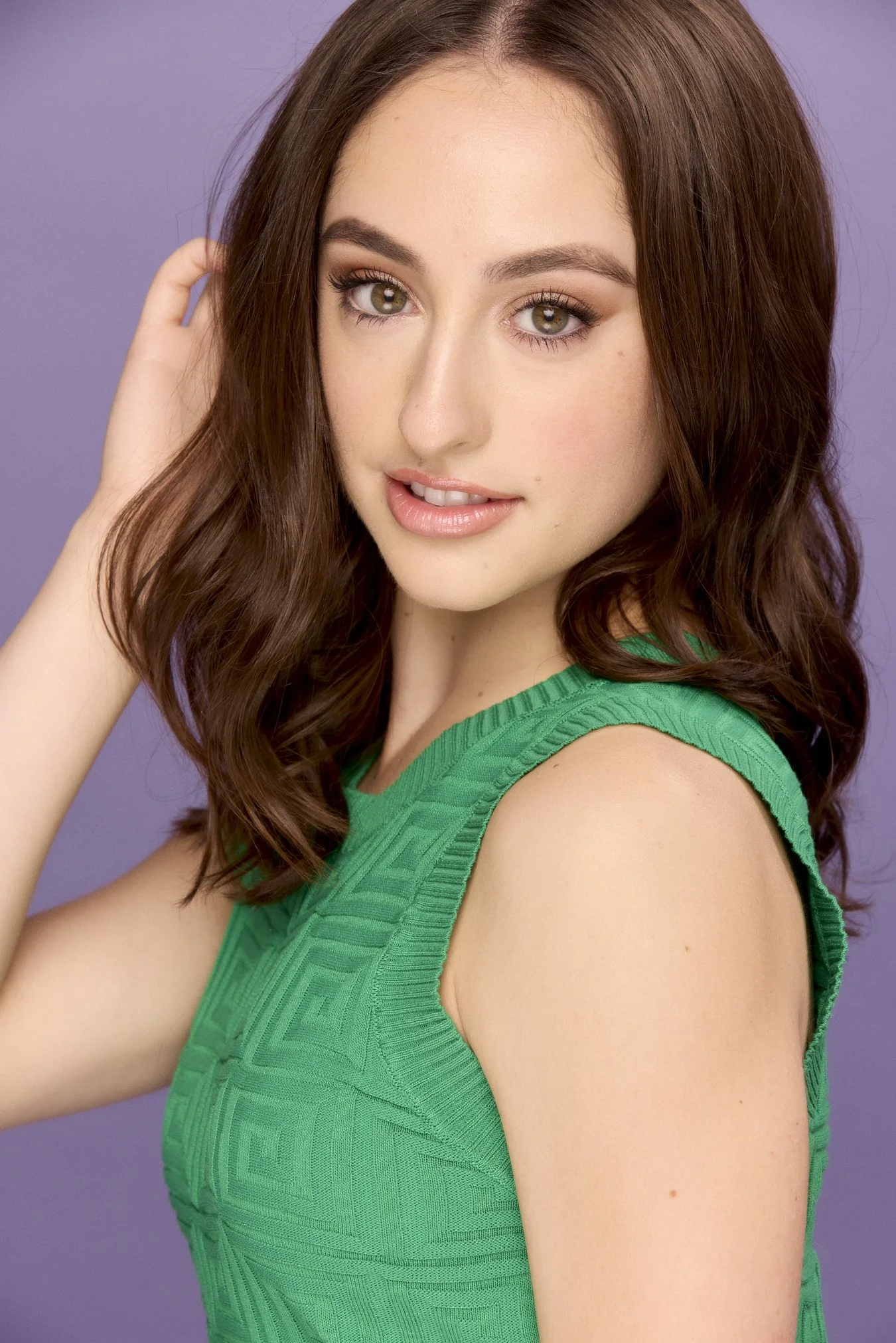 A young woman with long, wavy brown hair, green eyes, and light skin, wearing a sleeveless green dress, posing against a purple background.