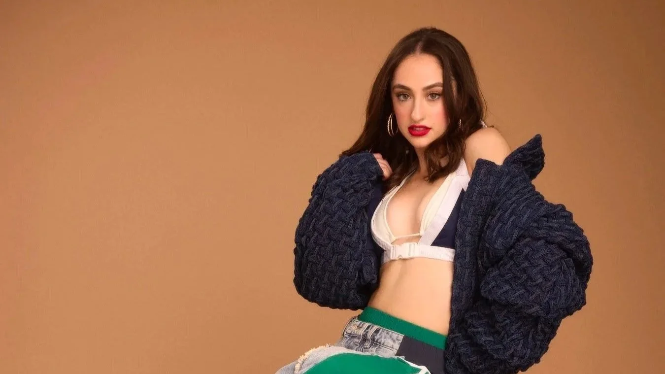 A young woman with dark hair and hoop earrings posing against a plain background, wearing a dark knitted cardigan, a beige sports bra, and multicolored pants.