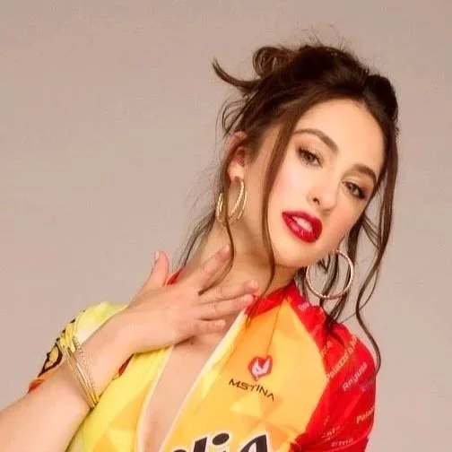 Young woman with curly hair, wearing large hoop earrings, red lipstick, and a colorful shirt, posing with her hand near her face against a plain background.