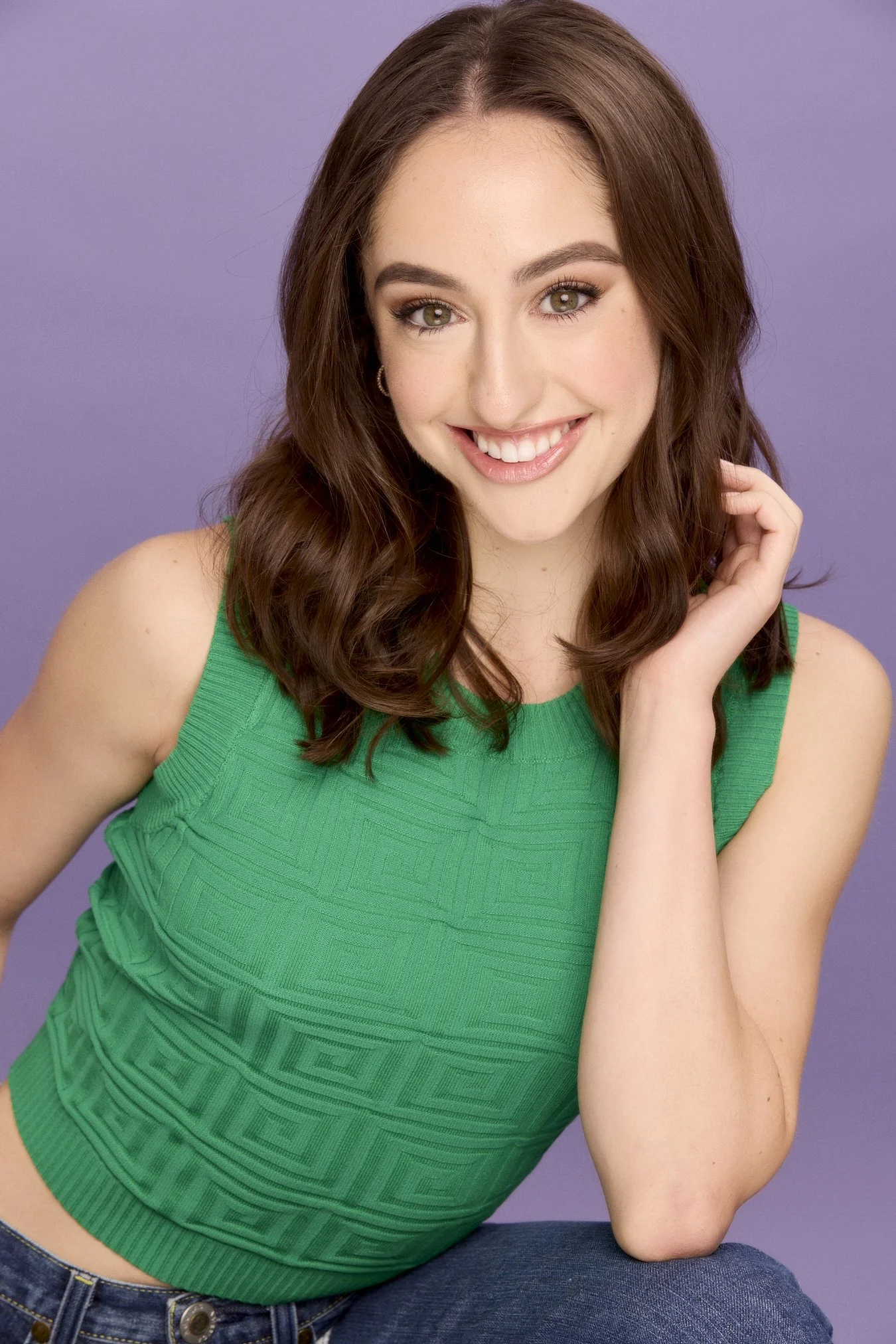 A young woman with long brown hair, green eyes, and light skin, smiling and posing with her hand near her face, wearing a sleeveless green top against a purple background.