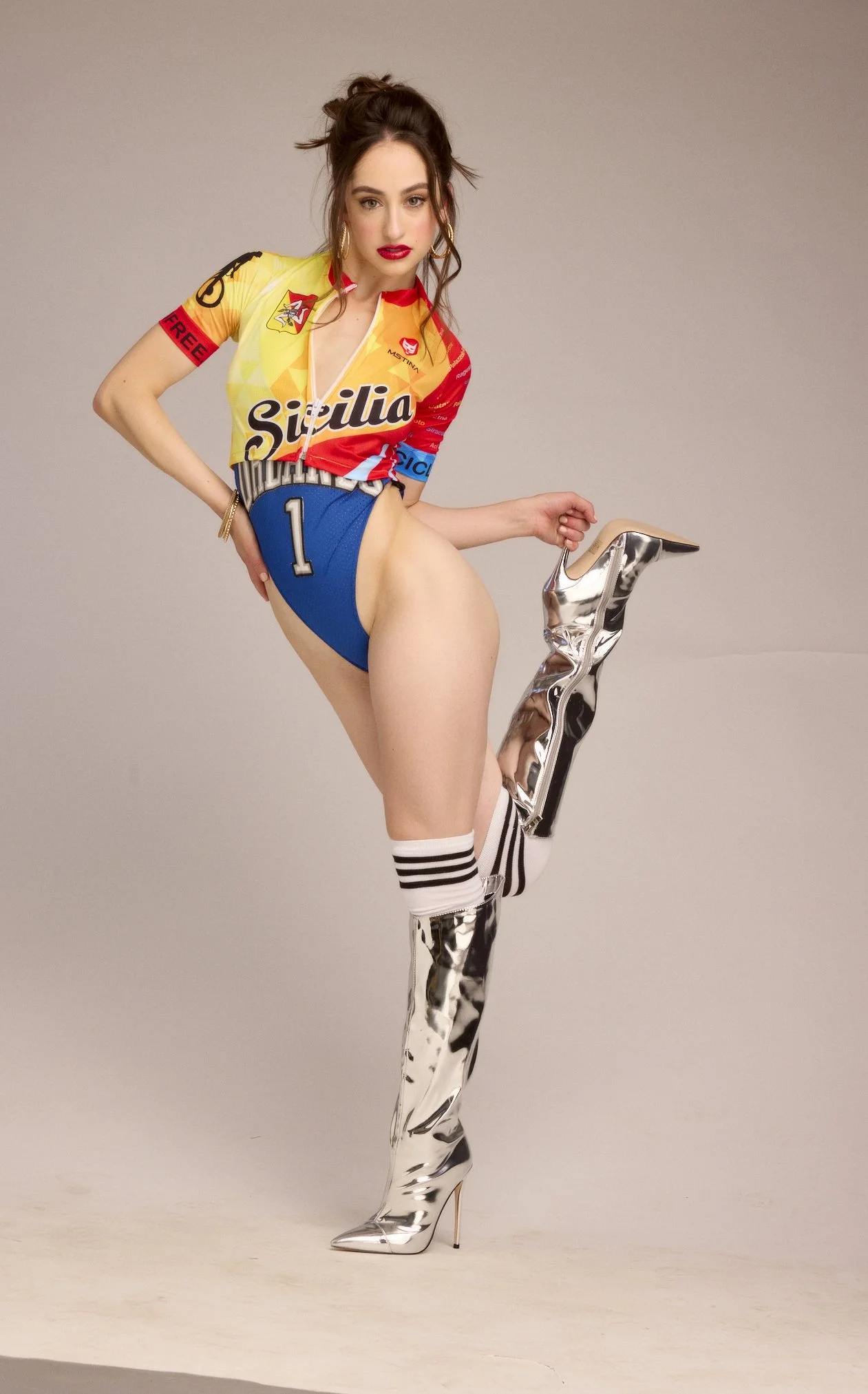 A woman with dark hair wearing a colorful racing jersey, blue shorts, knee-high striped socks, and shiny silver thigh-high boots posing with one leg raised.