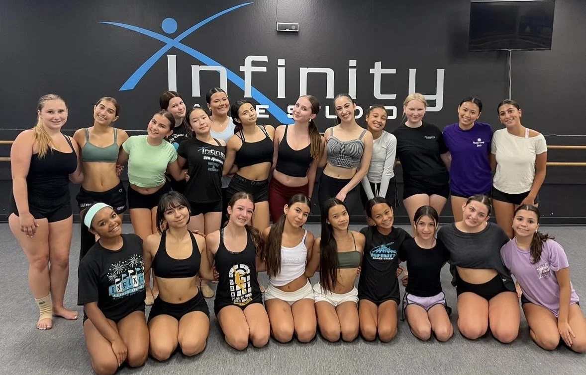 Group of young women in workout clothes posing in a dance studio with the Infinity Dance Studio logo on the wall.