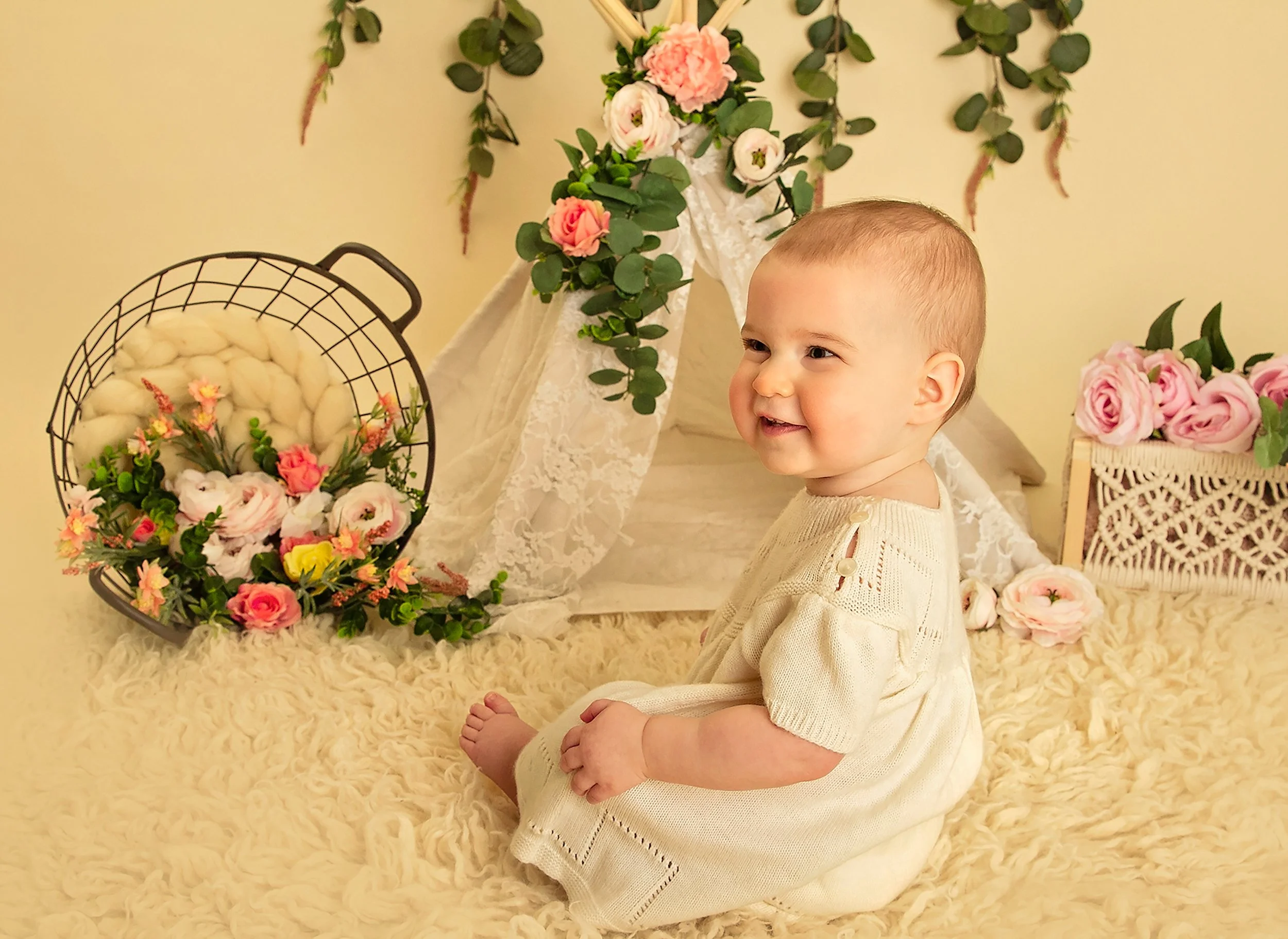 A young child sitting on a fluffy cream-colored rug in front of a floral display, including a basket of pink and yellow flowers, with roses and greenery in the background.